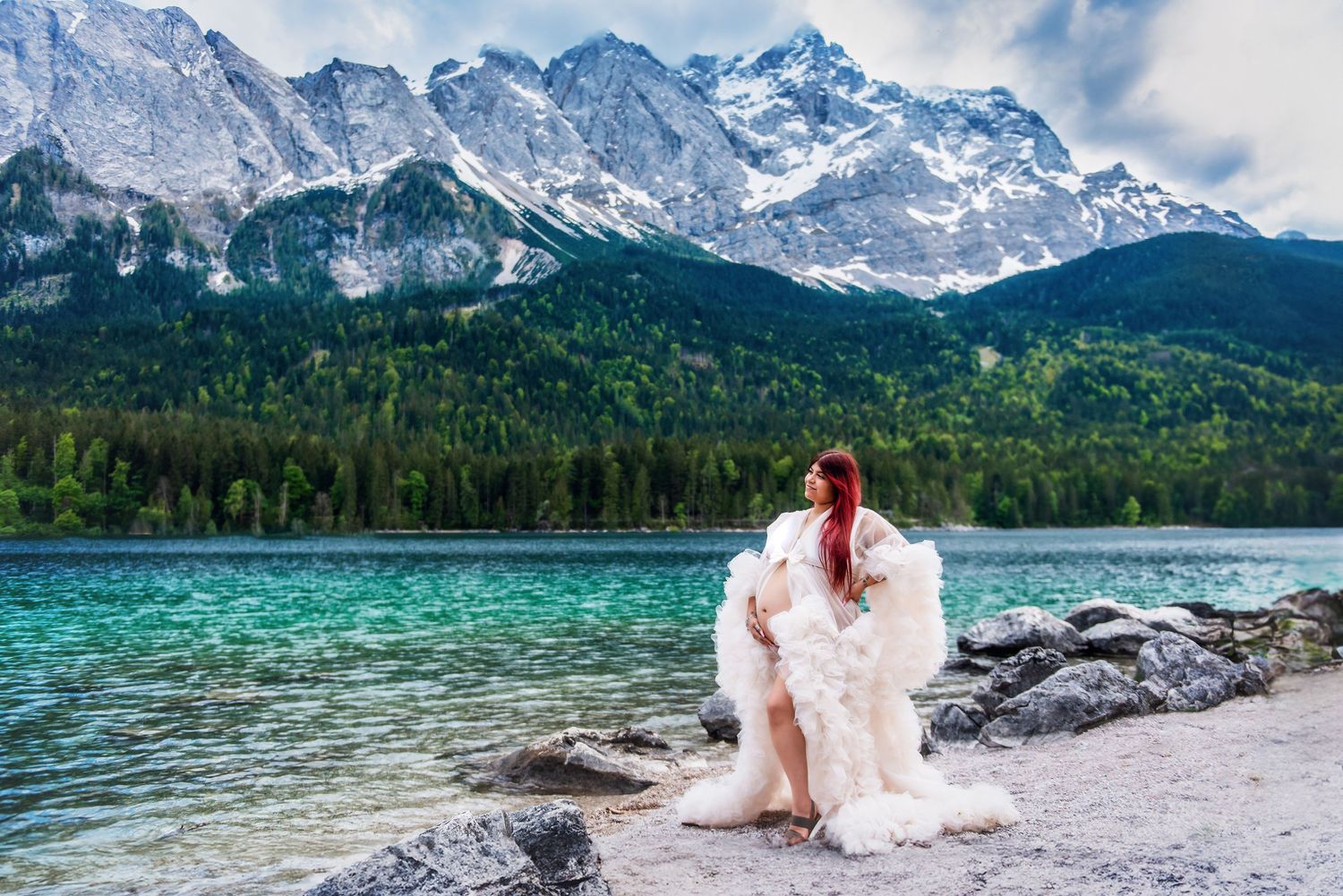 Someone in a flowing white dress sits on rocks beside a turquoise alpine lake with snow-capped mountains in the background.