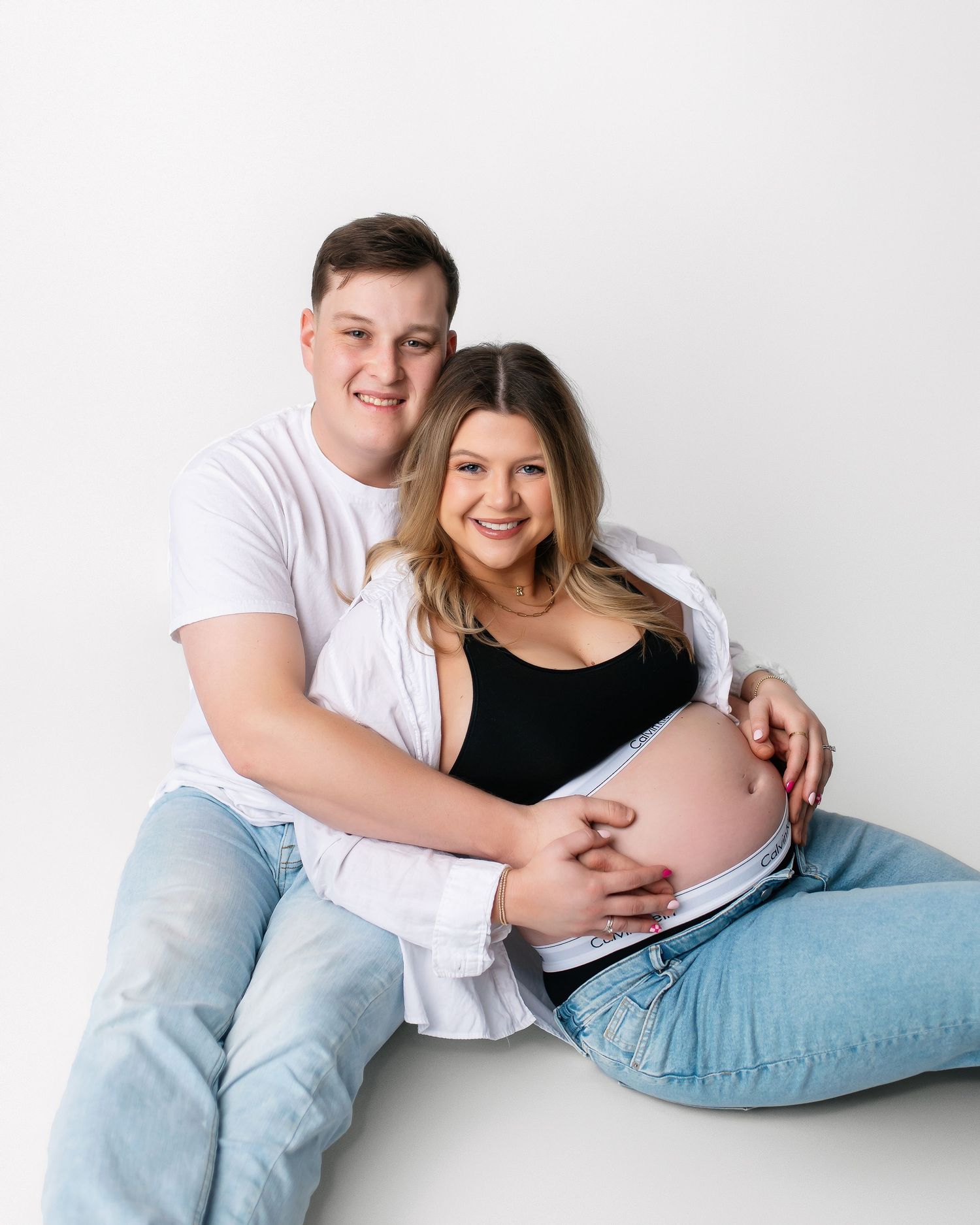 Couple sits together on floor during casual maternity photoshoot in studio setting.