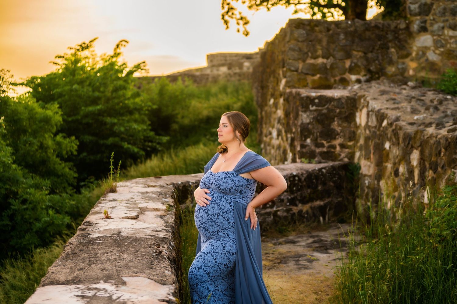 Pregnant woman in blue dress poses against stone ruins during sunset photoshoot.