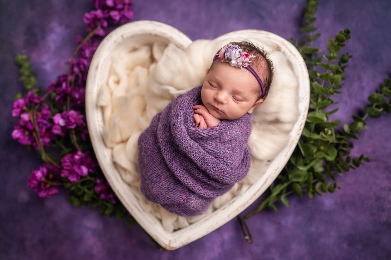 Newborn wrapped in purple swaddle sleeping in heart-shaped bowl with purple flowers.