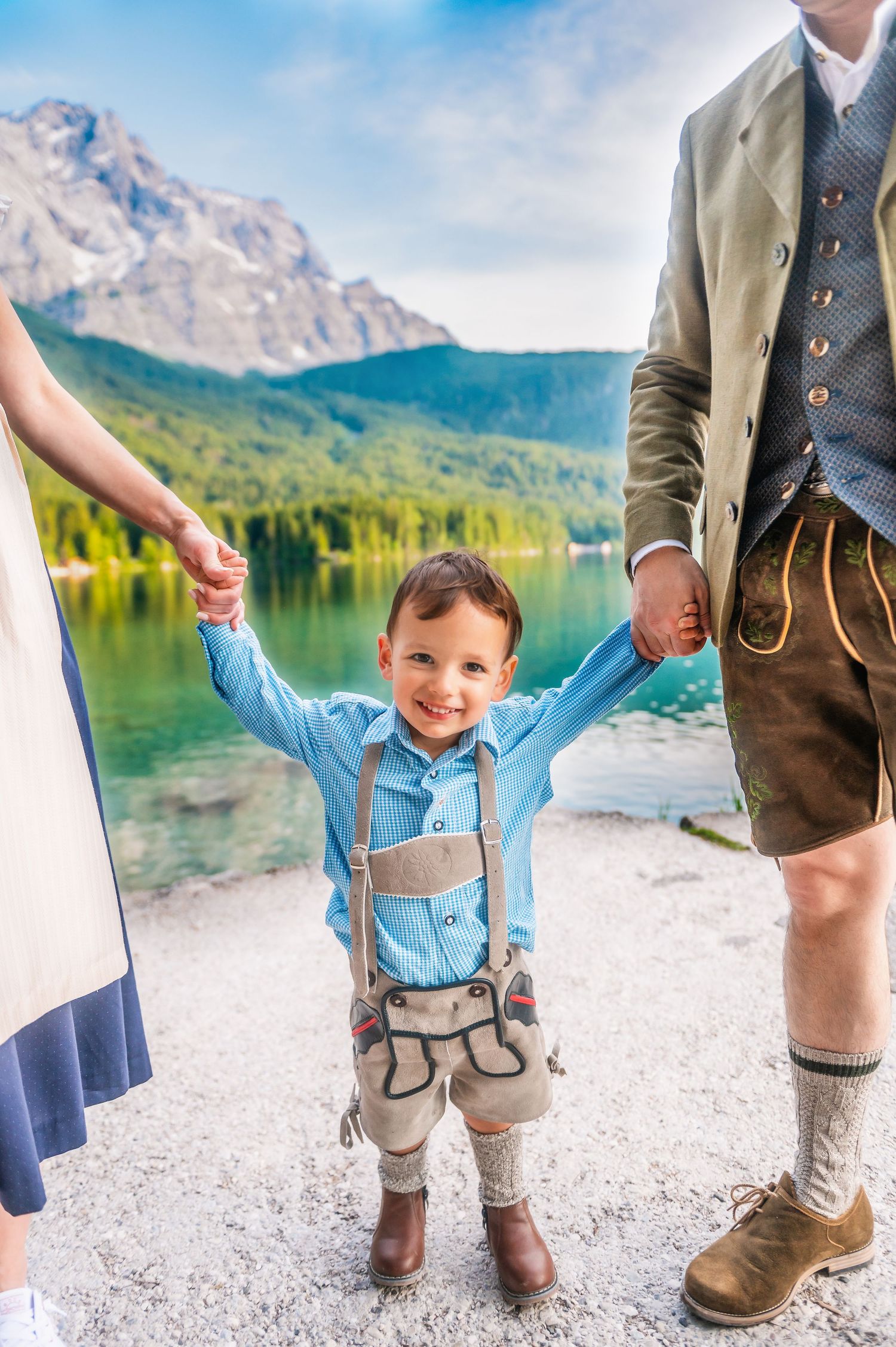 Young child wearing traditional Bavarian lederhosen and blue shirt stands by alpine lake with mountain backdrop.