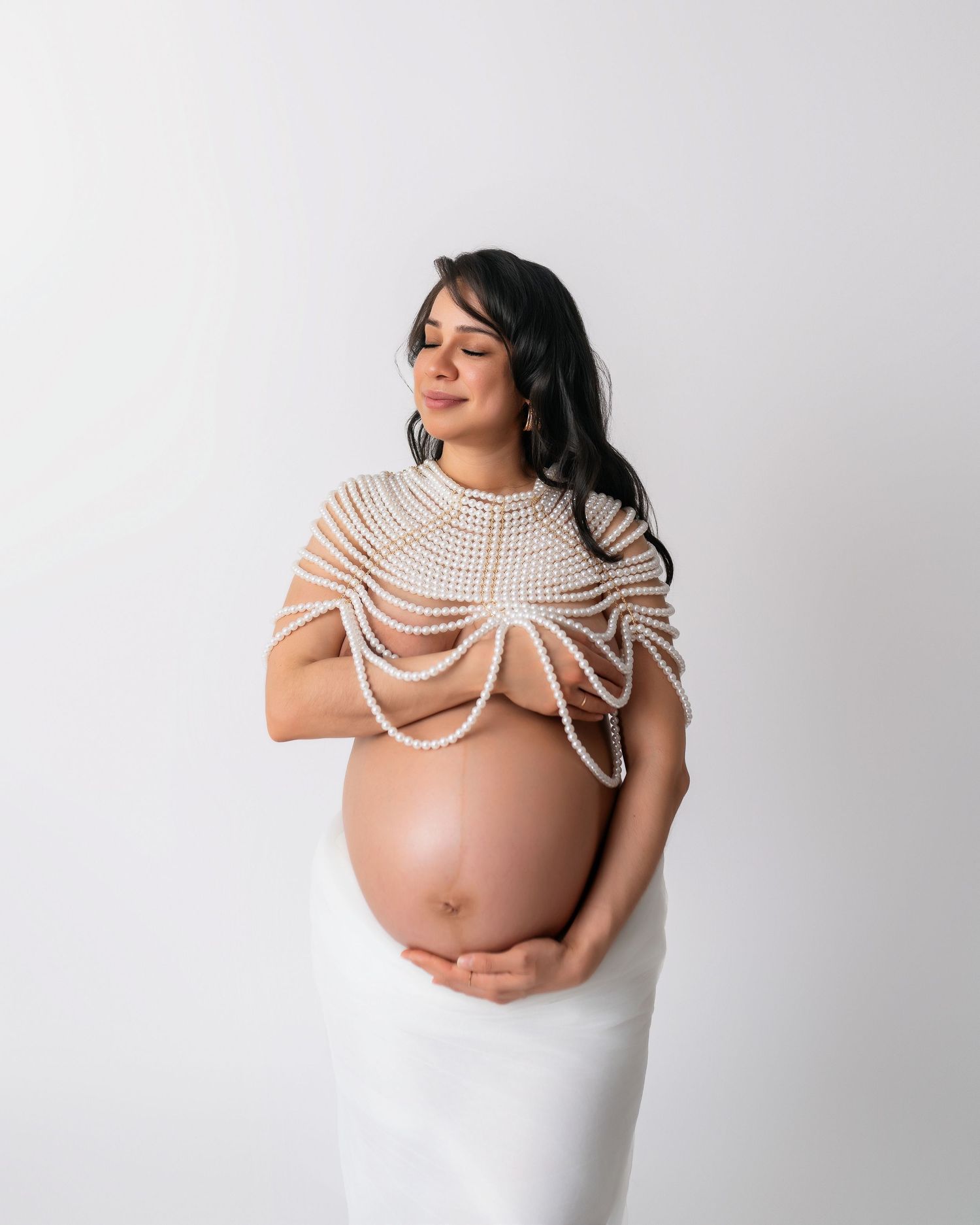 Elegant maternity portrait with woman wearing white dress and draped pearls.