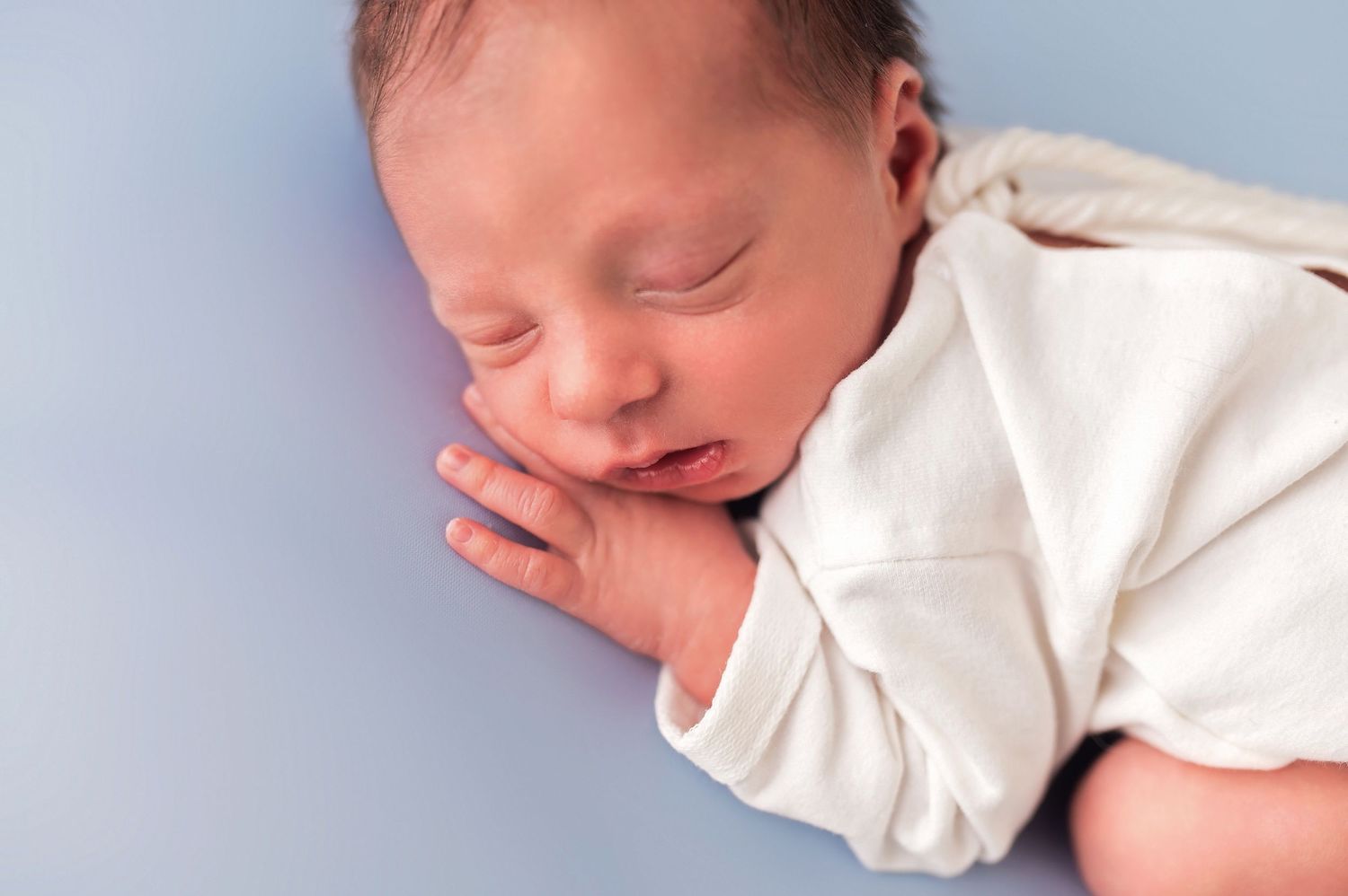 Newborn baby sleeping peacefully on light blue background with matching wrap and headband pillow.