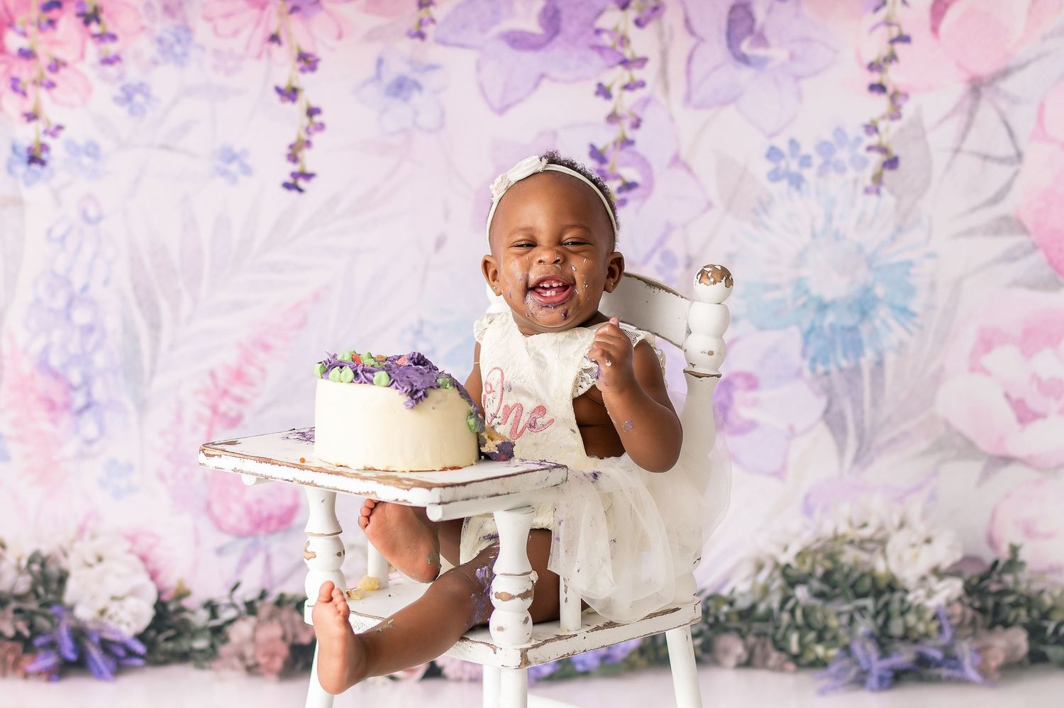 Cake smash photo session with dreamy pink and purple balloon backdrop and white decorations.