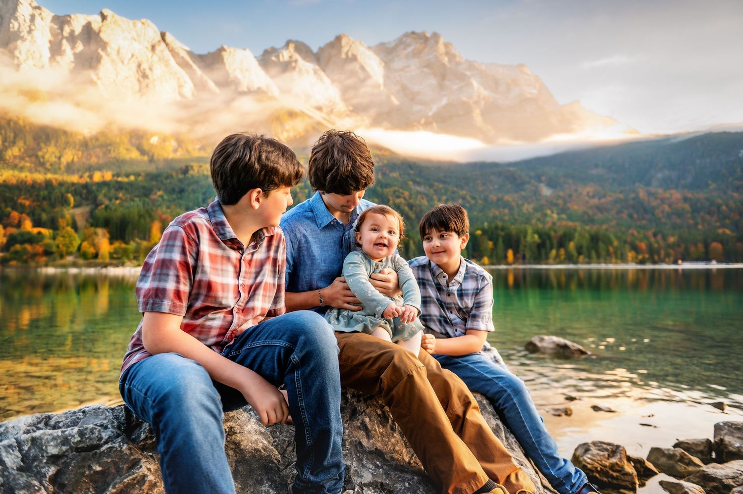 Family enjoying a peaceful moment together on a rocky lakeshore with mountains and forest in the background.