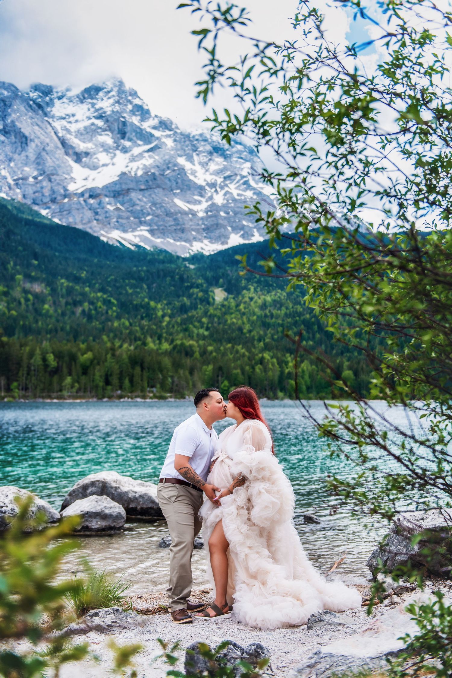 A couple shares a romantic moment by a pristine alpine lake surrounded by snow-capped mountains and lush forest.