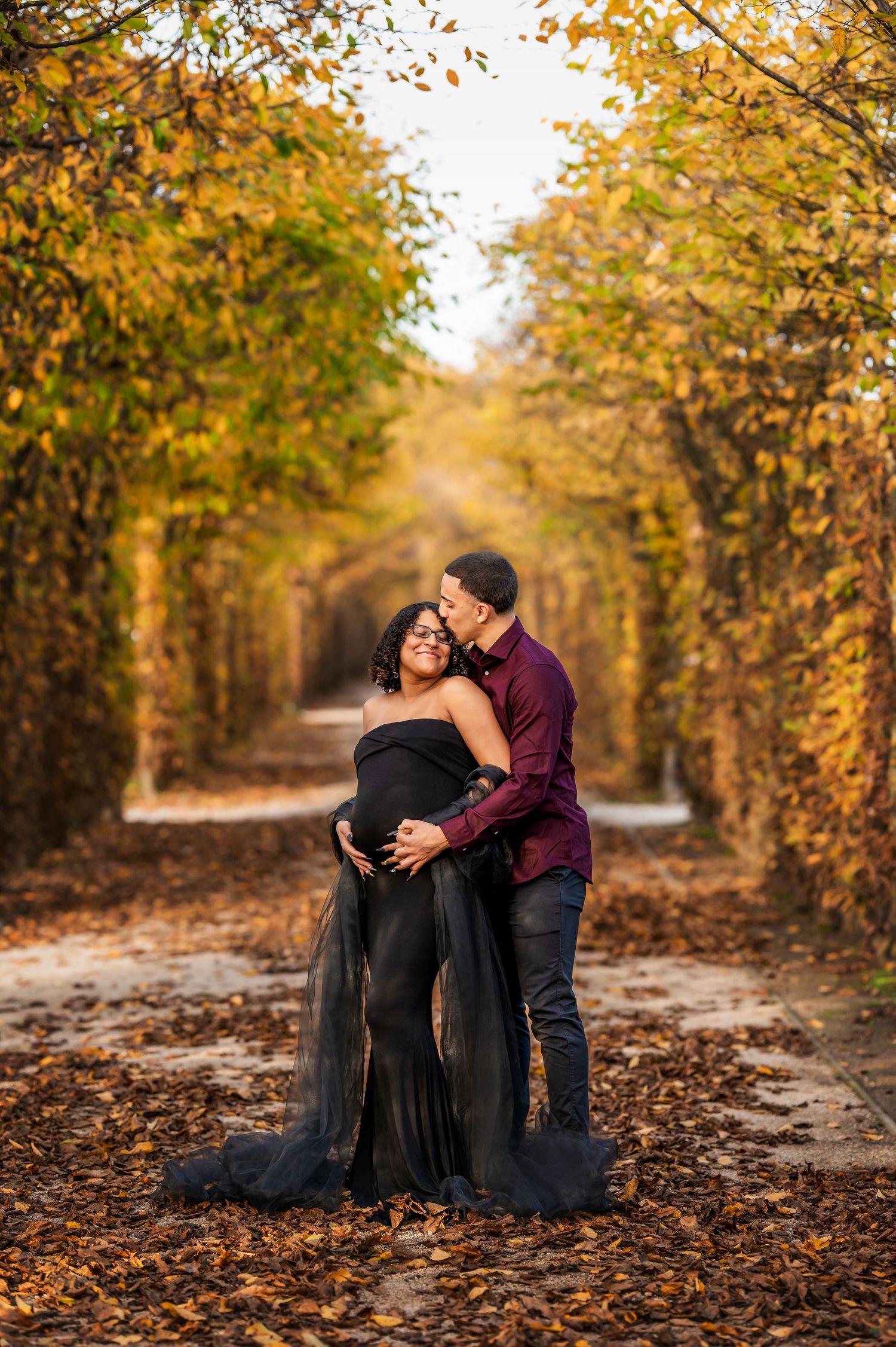 Intimate maternity moment captured in tree-lined autumn path.
