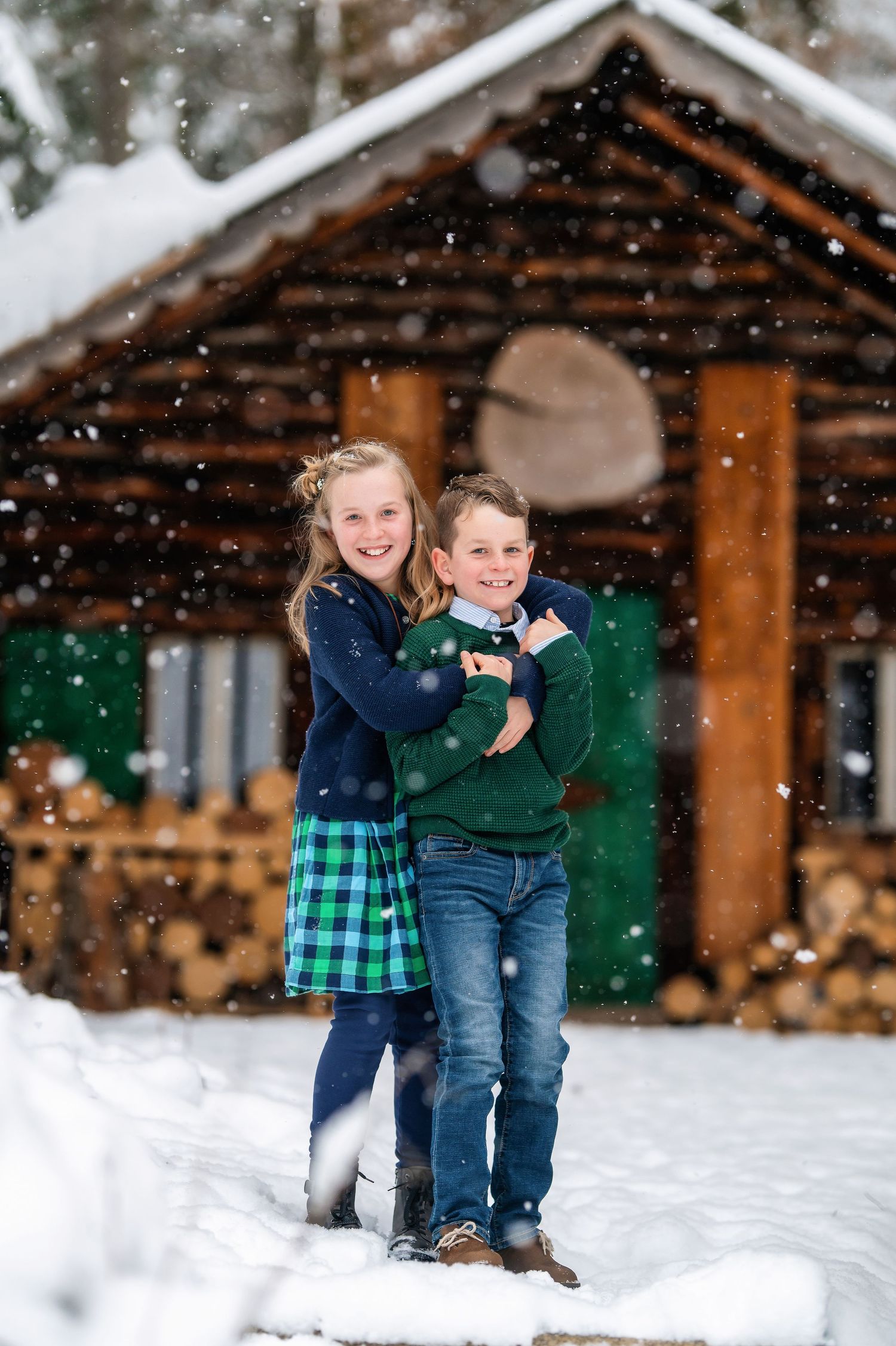 Two children play and smile together in front of a snow-covered cabin.