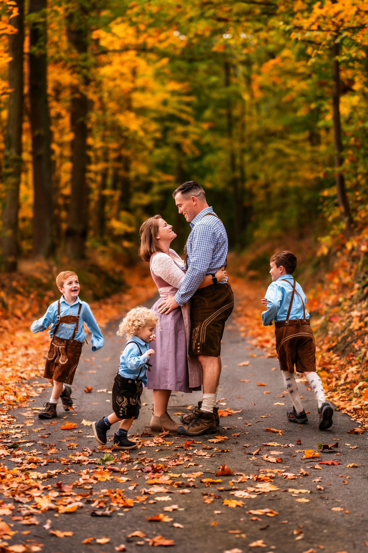 Family in traditional German outfits walks along autumn forest path covered in fallen leaves.