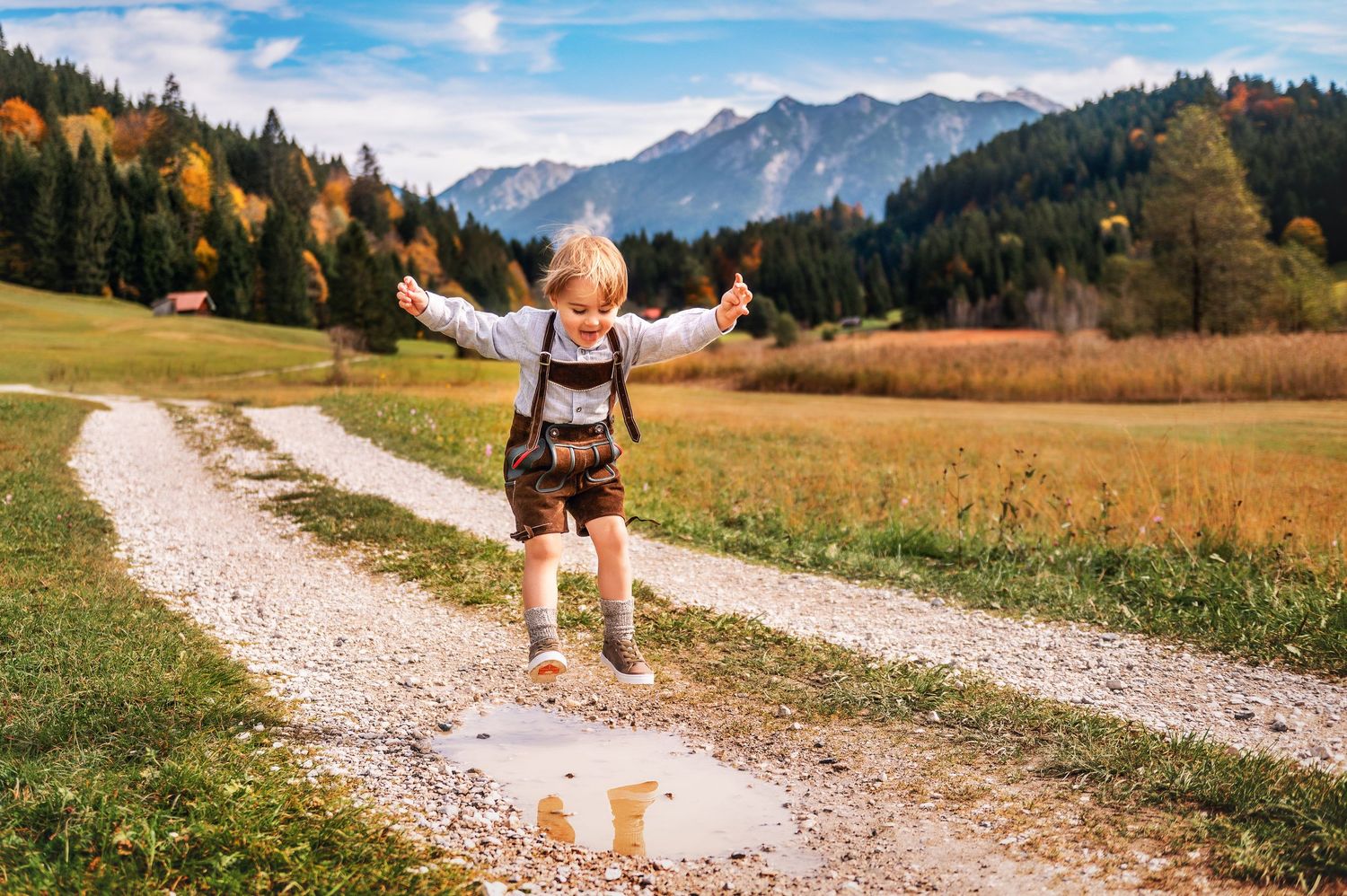 A hiker in traditional Bavarian lederhosen walks along a scenic mountain trail surrounded by autumn foliage in the Alps.