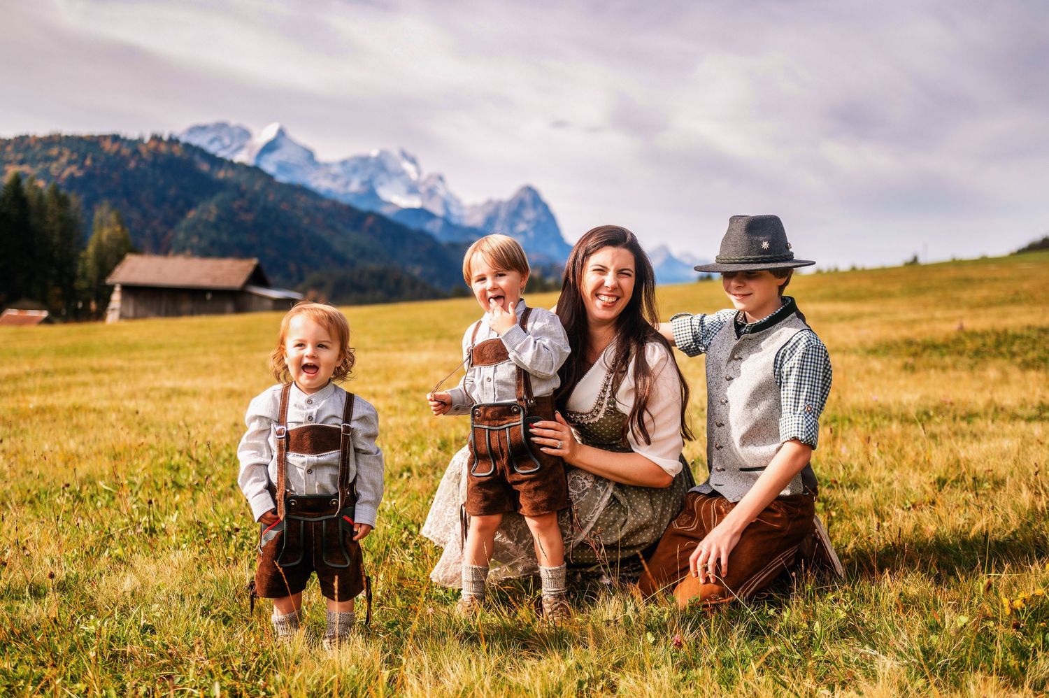A family wearing traditional Alpine clothing poses together in a meadow with mountains and a rustic cabin in the background.