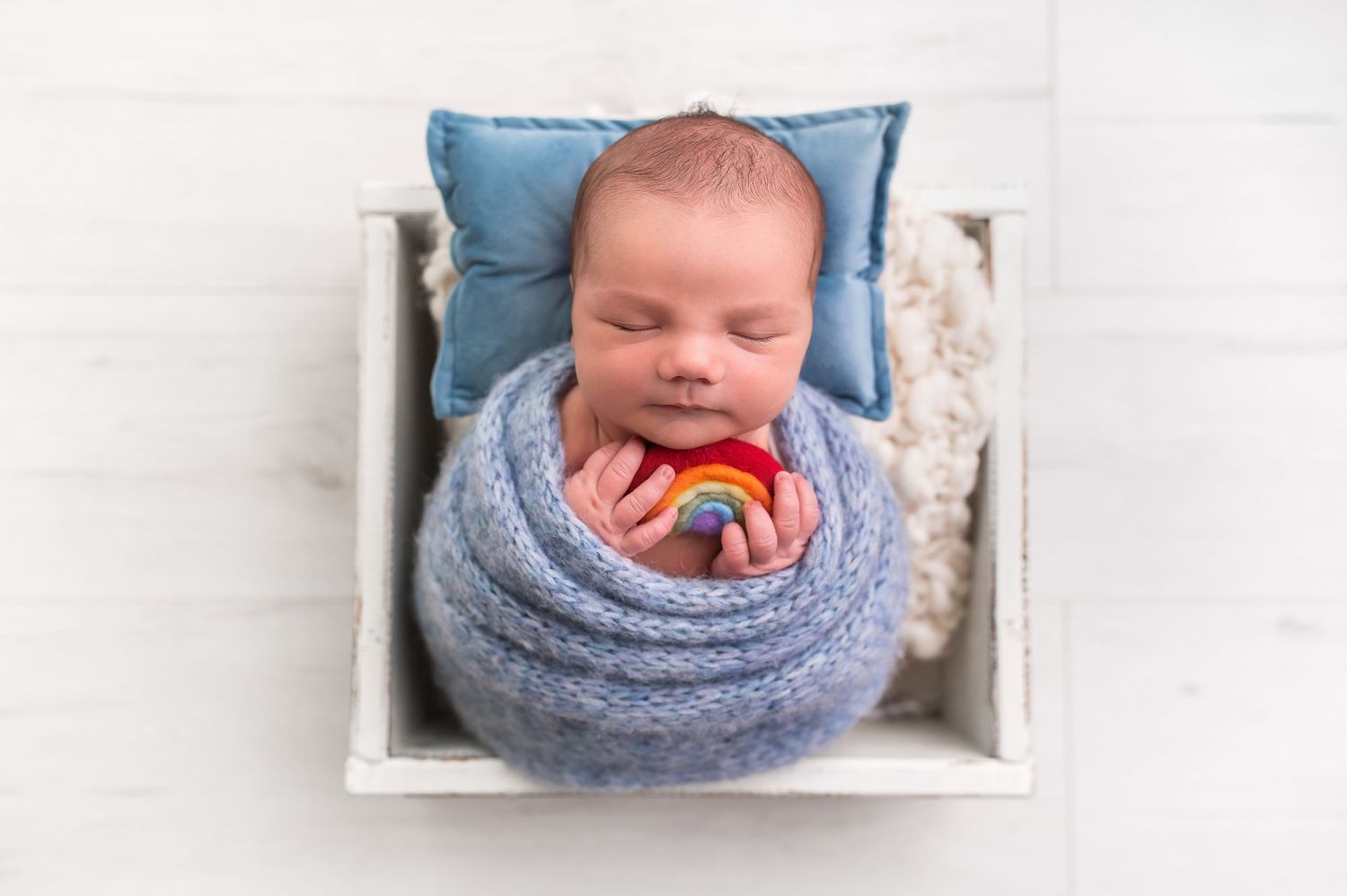 Newborn baby wrapped in blue blanket lying in white wooden prop bed.