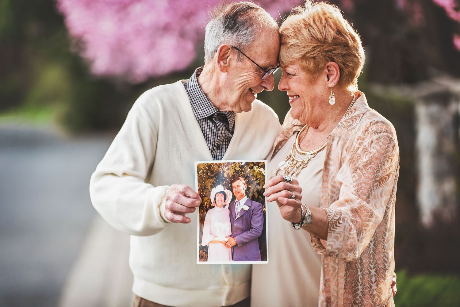 Elderly couple smiling and holding their old wedding photo outdoors with pink blossoming trees in the background.