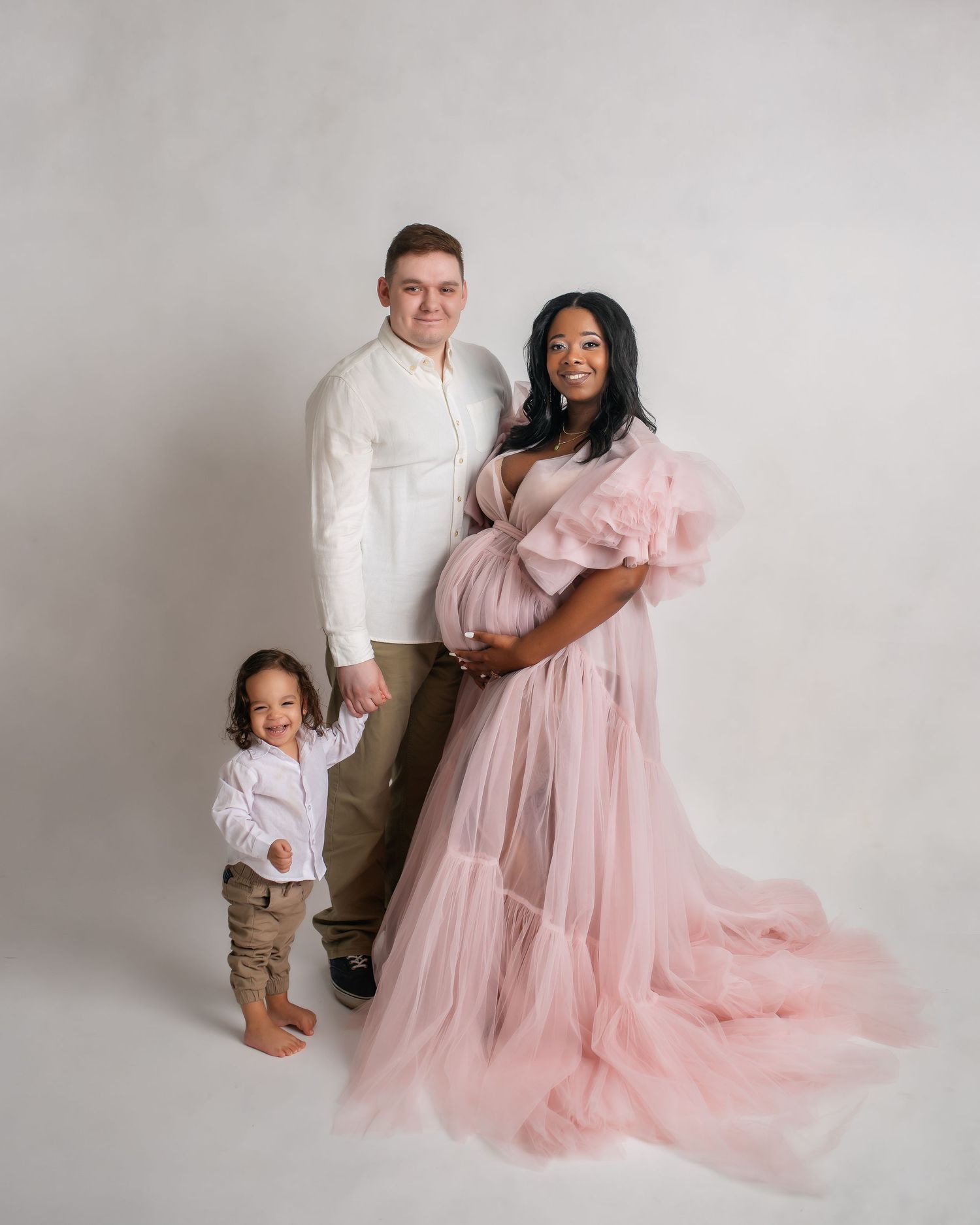 Family maternity photo featuring a pink tulle gown against white backdrop with toddler in beige pants.