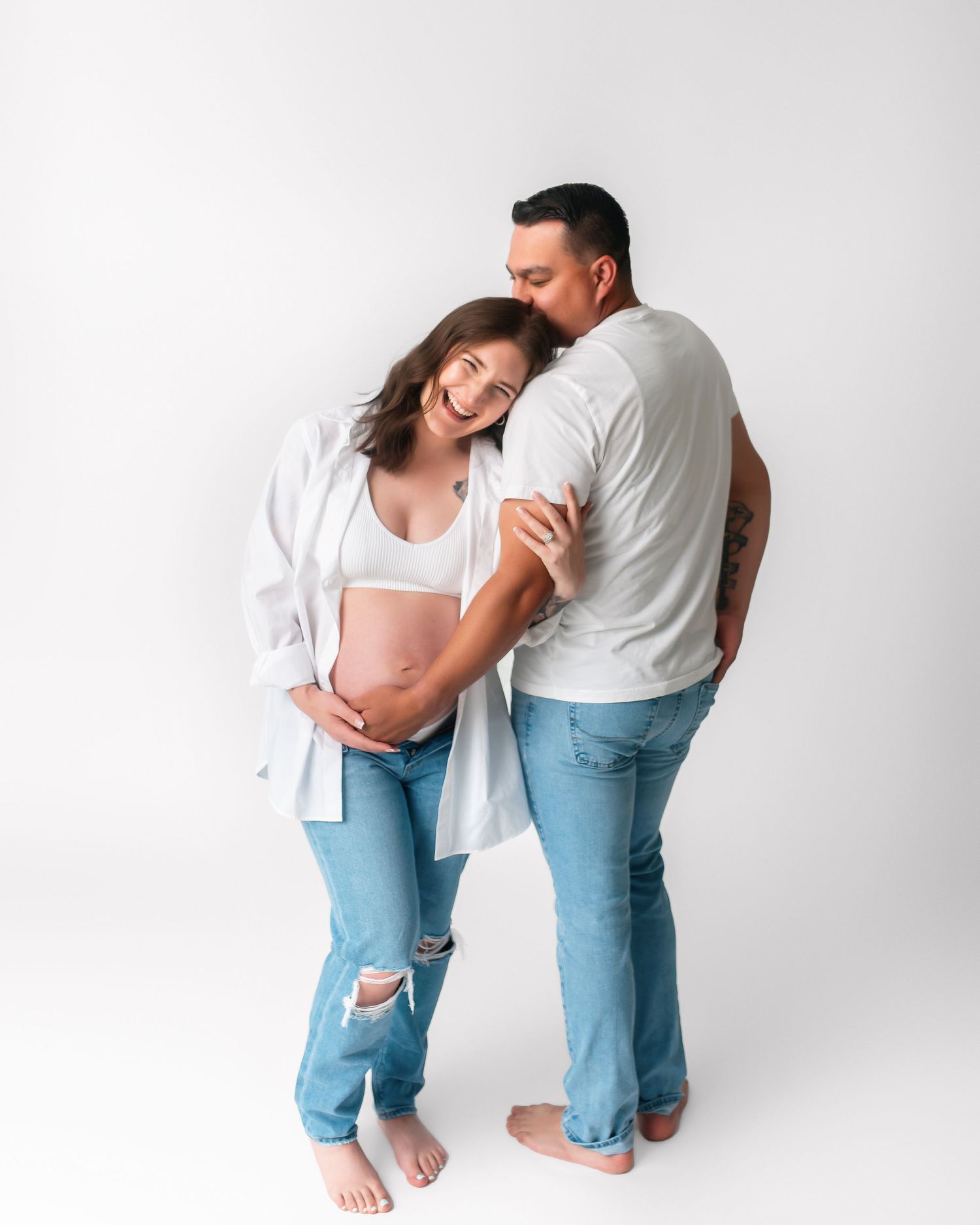 Tender maternity photo of couple in white and denim against studio backdrop.