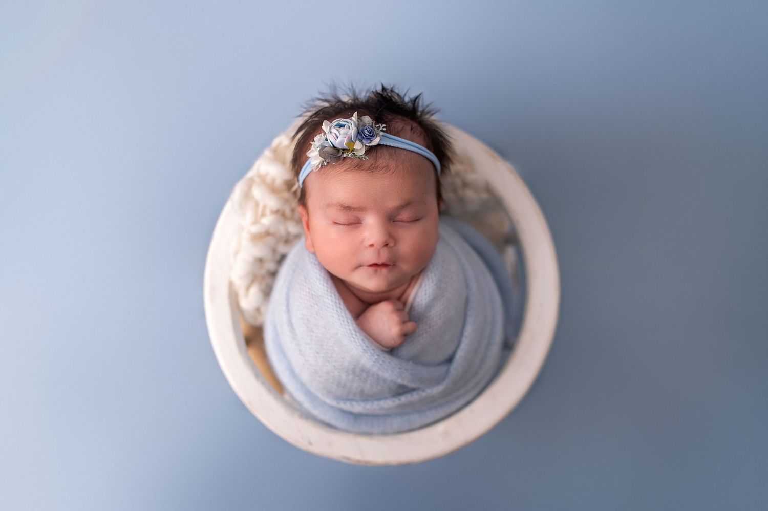 Professional newborn portrait with baby in gray wrap against light blue backdrop.