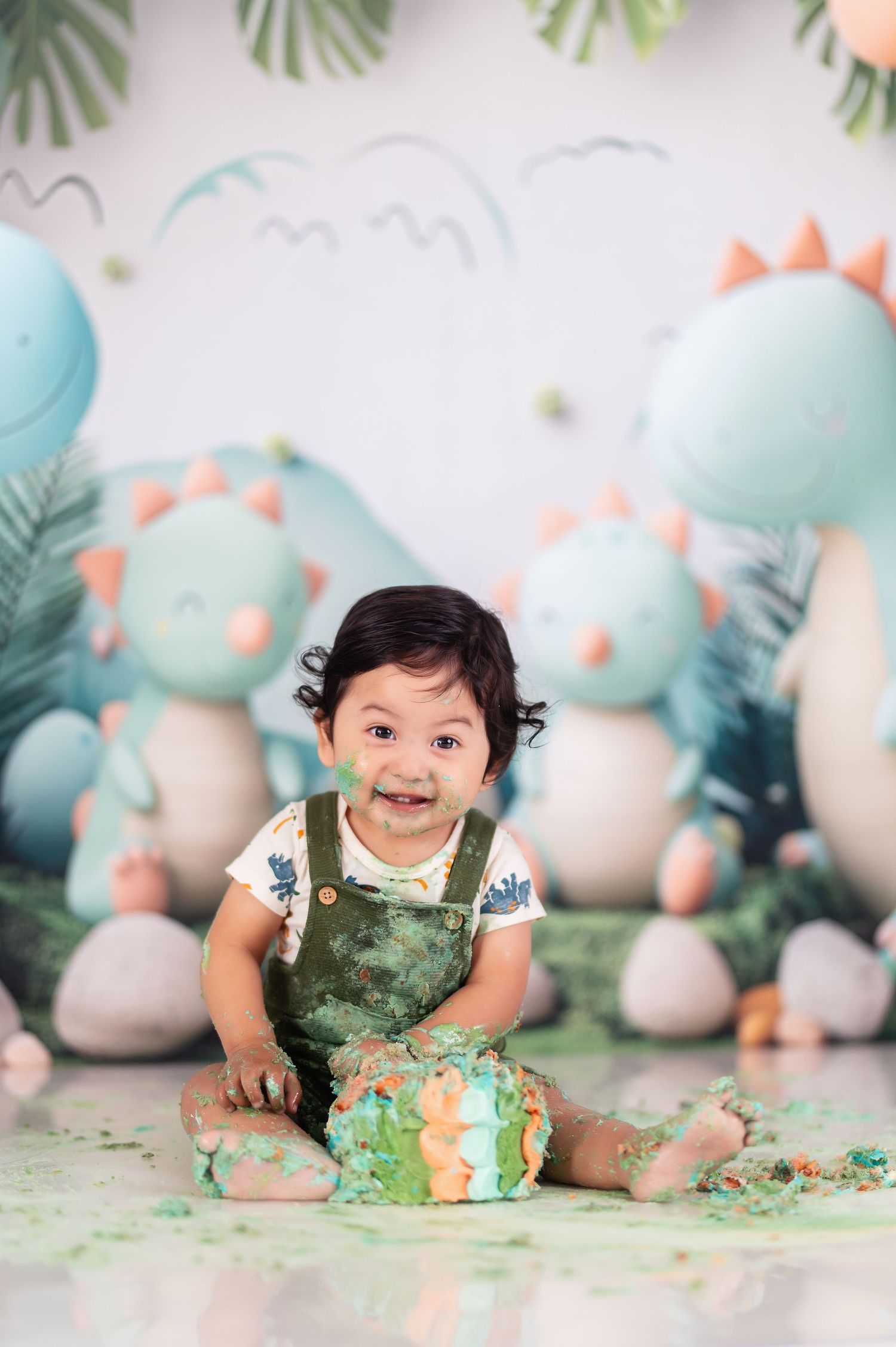Baby wearing white shirt and green overalls during dinosaur themed cake smash session.