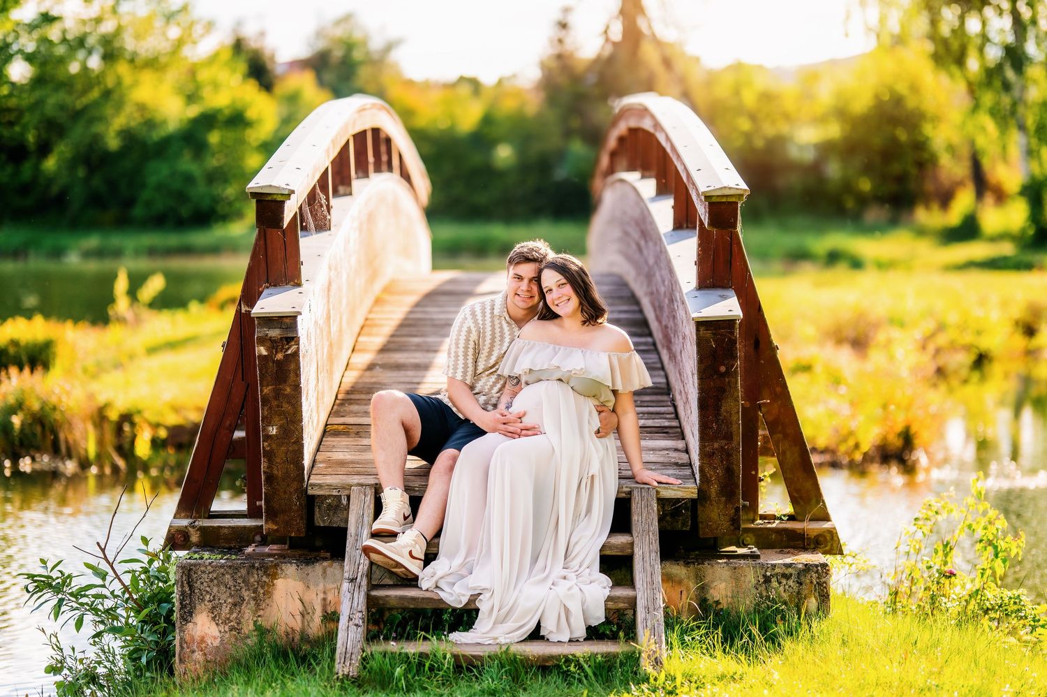 Maternity portraits on wooden bench surrounded by golden sunlit meadow.
