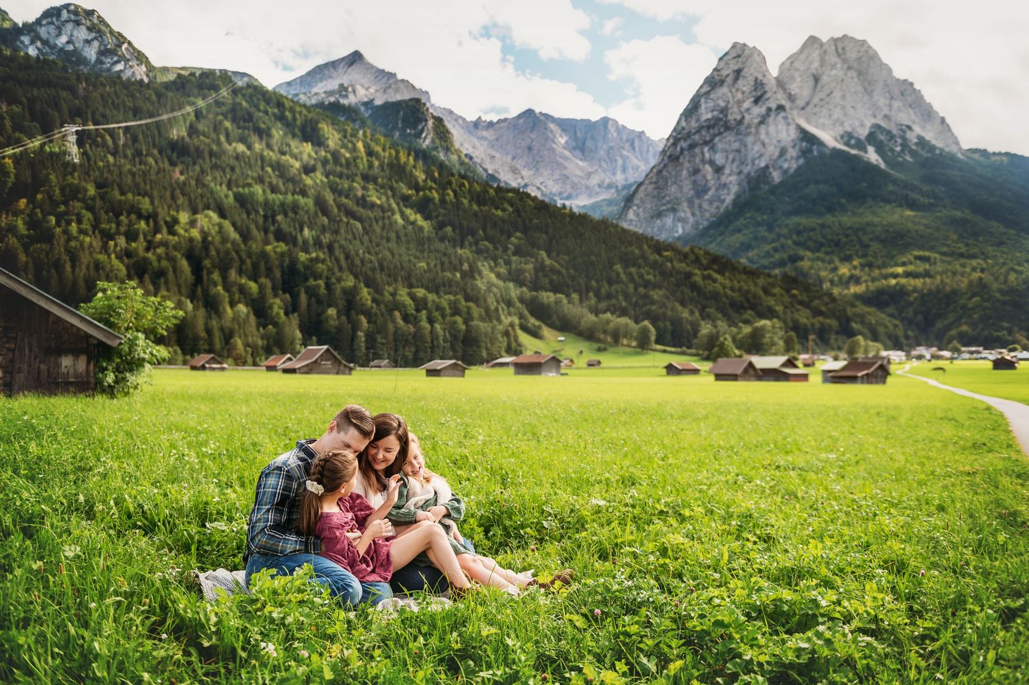 Couple sitting together on lush green meadow with Alpine mountains and traditional wooden chalets in background.
