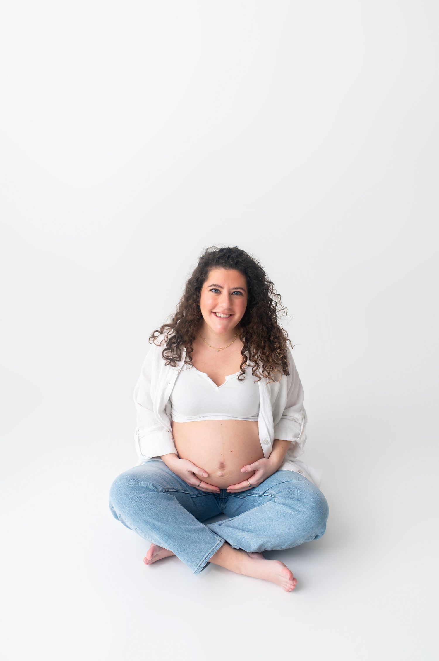 Pregnant woman in white shirt and jeans sits cross-legged on white backdrop during maternity shoot.