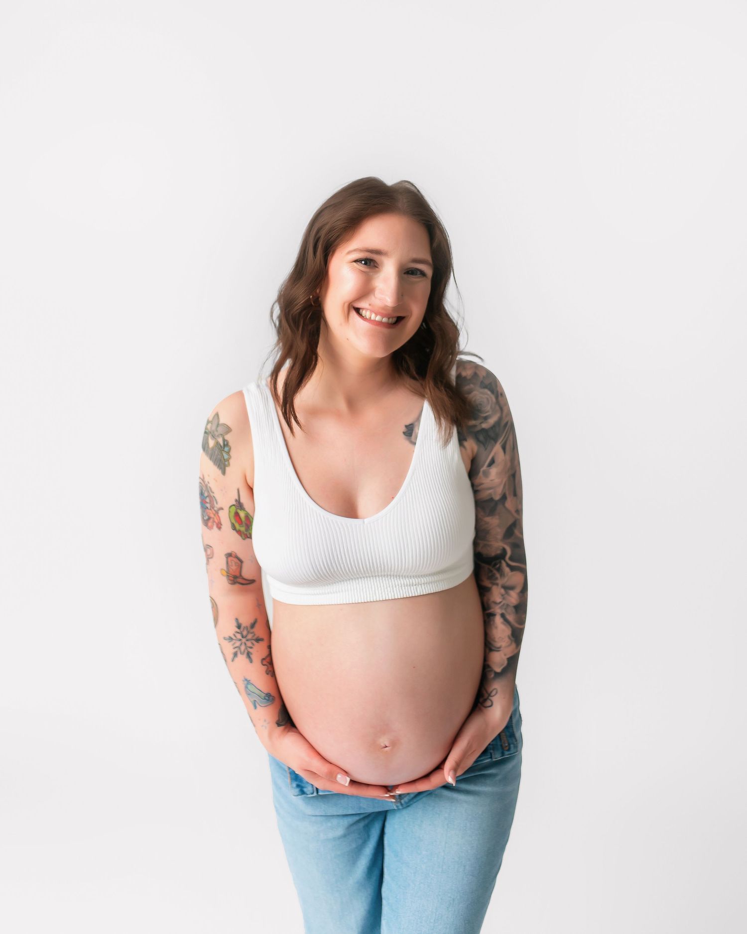 Smiling pregnant woman with sleeve tattoos poses in casual attire against white background.