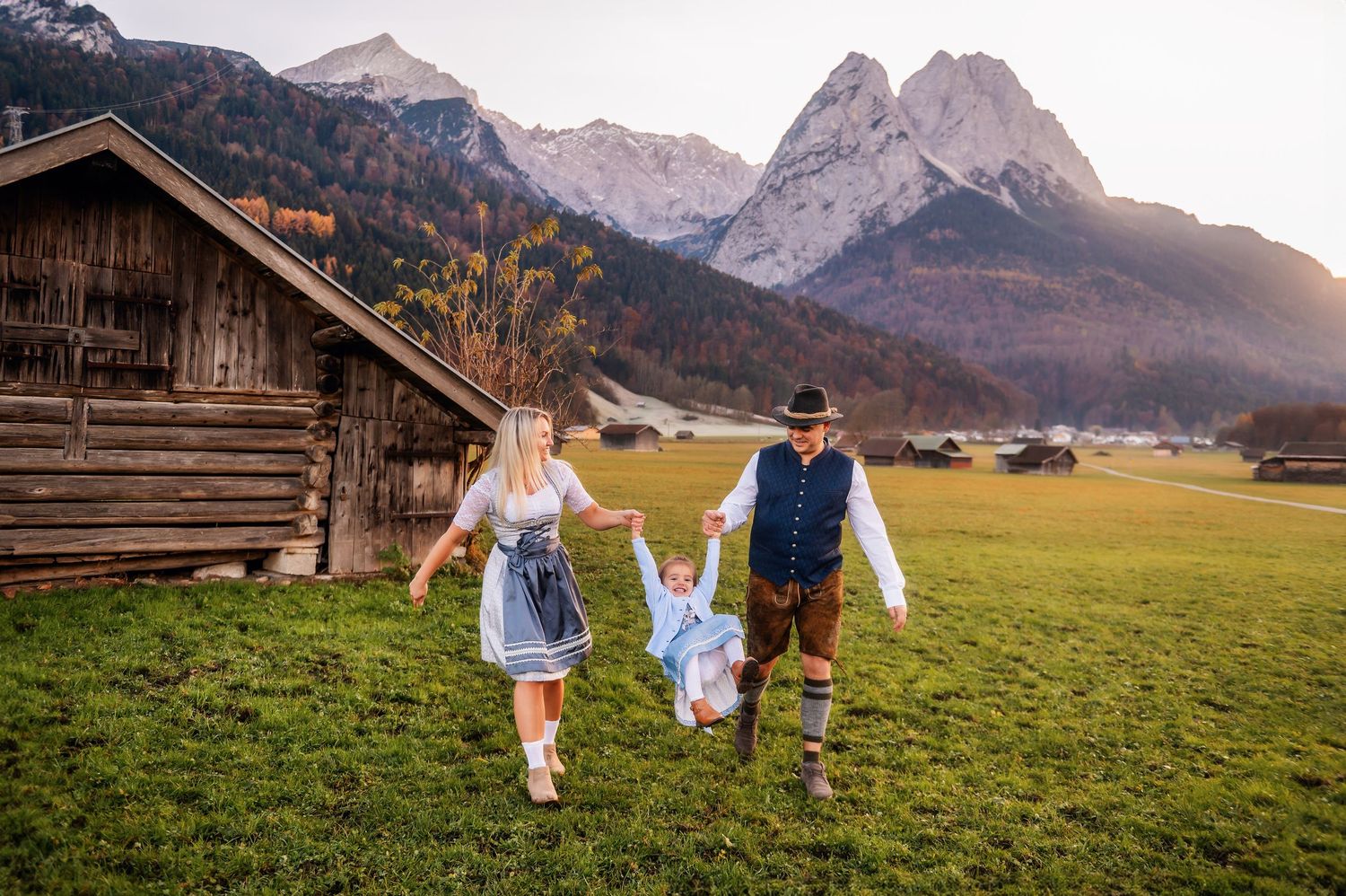 Traditional Bavarian scene with people in dirndl and lederhosen walking across alpine meadow with mountain peaks at sunset.