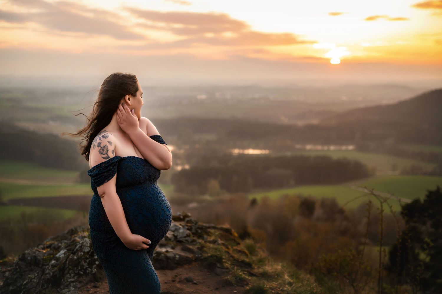 A silhouette stands at a mountain overlook during golden sunset with rolling hills and countryside below.