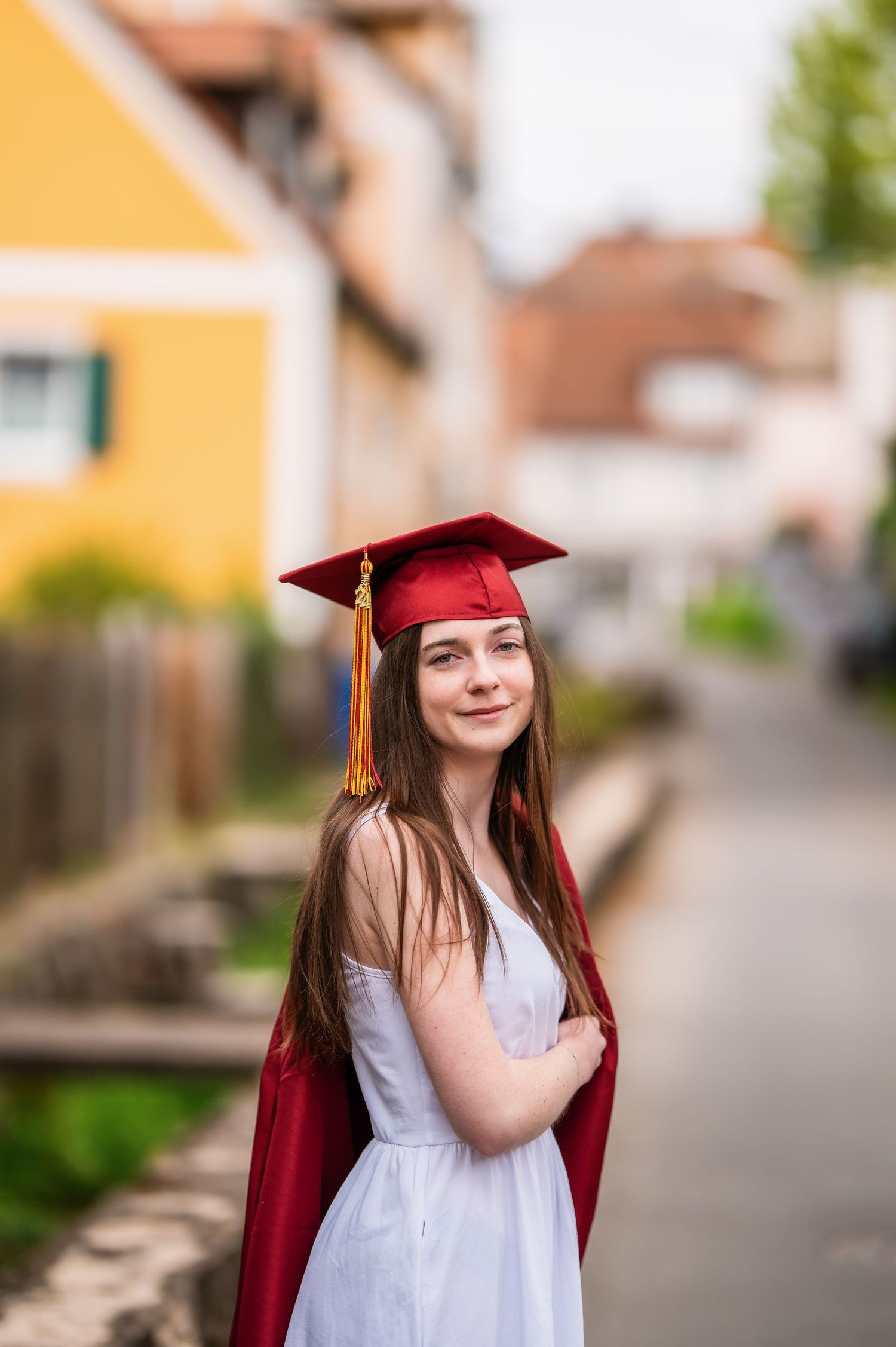 Graduate in red cap and gown poses on a street with yellow buildings in background.