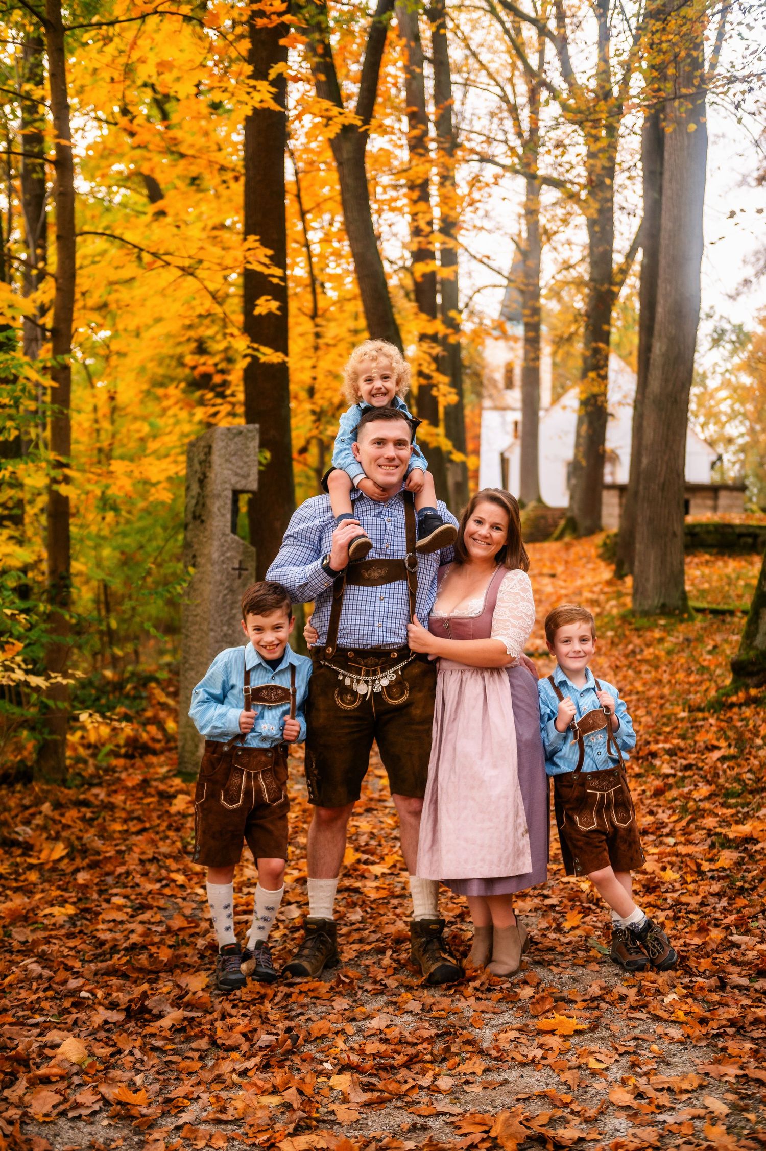 Family wearing traditional German attire poses among vibrant fall foliage with a church spire visible.