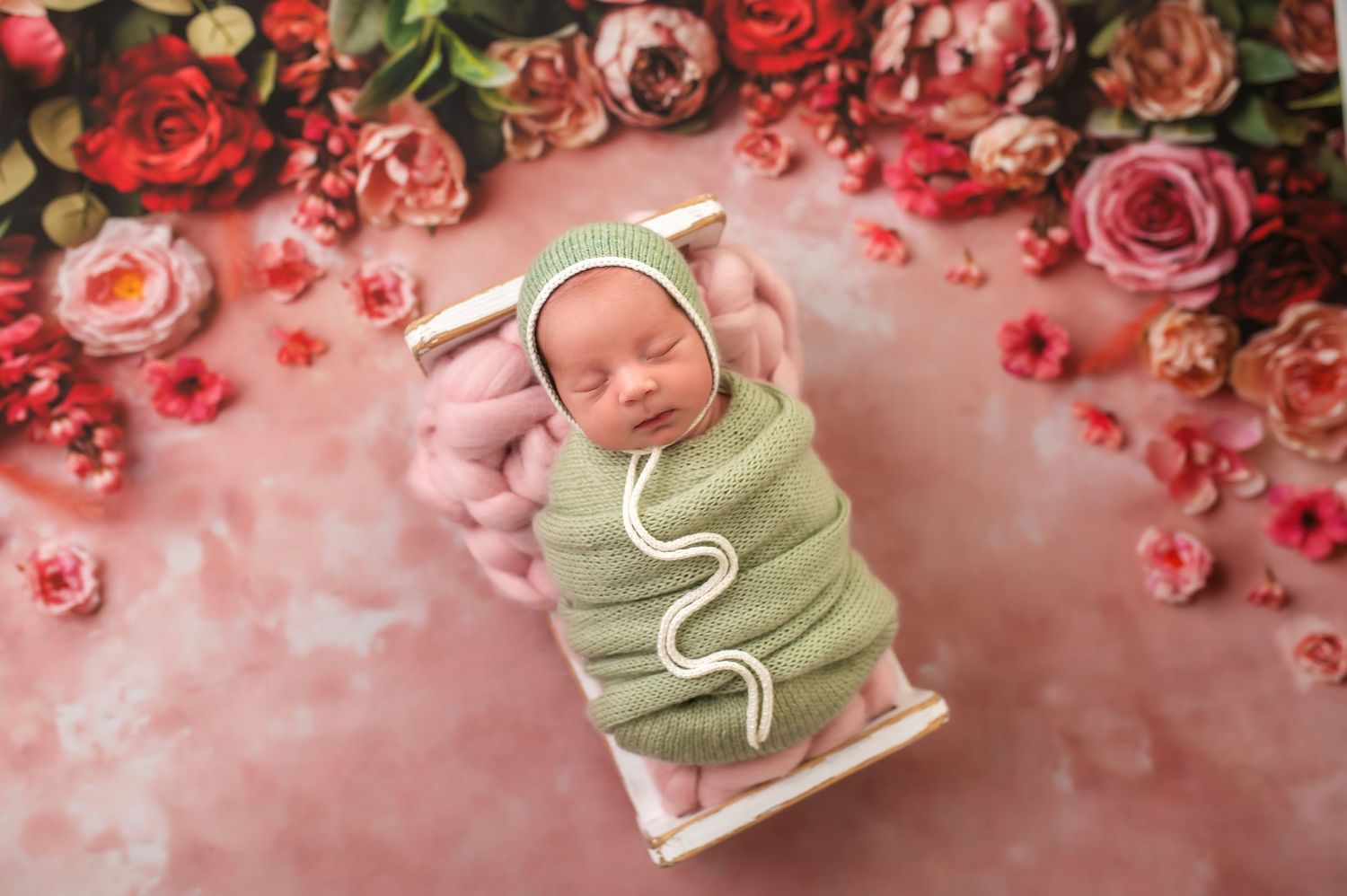 Sleeping newborn in mint wrap on small bed surrounded by pink and red roses.