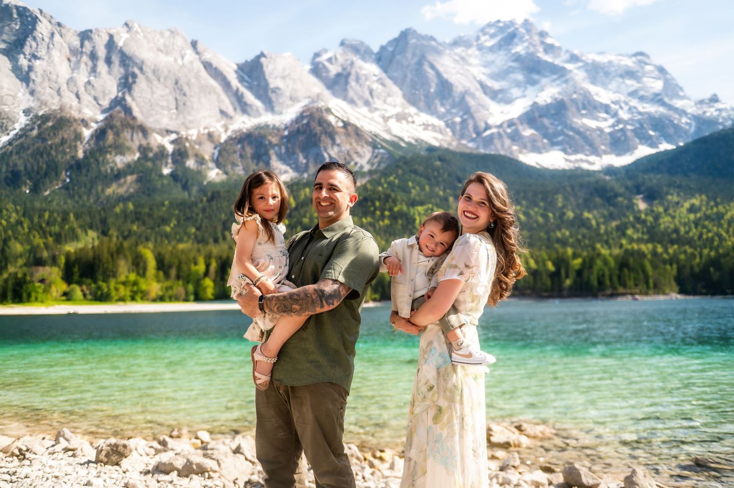 Family of four smiling by a turquoise alpine lake with snow-capped mountains and pine forests in the background.