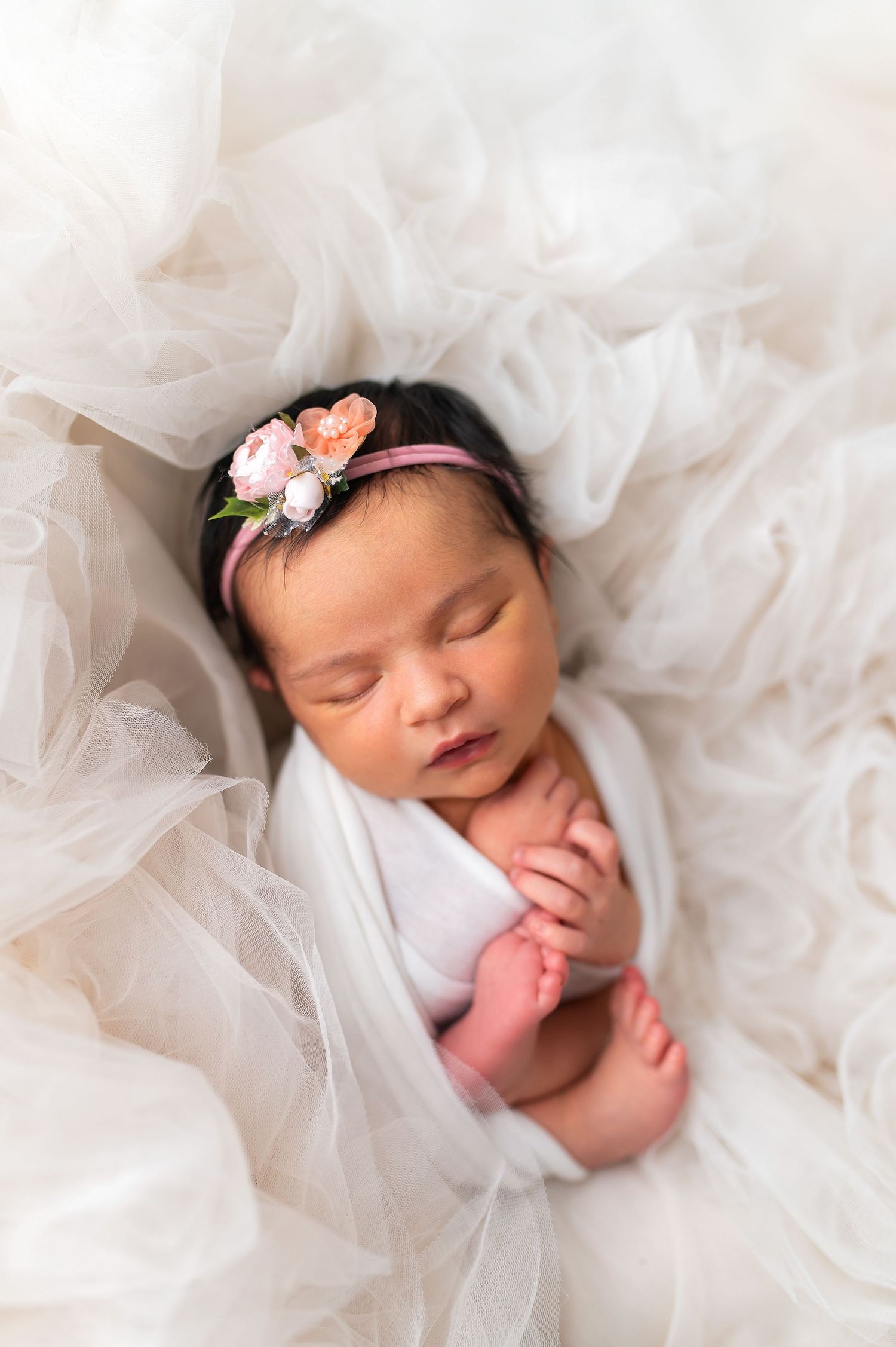 Newborn baby sleeping peacefully on soft white tulle fabric with delicate pink floral headband.