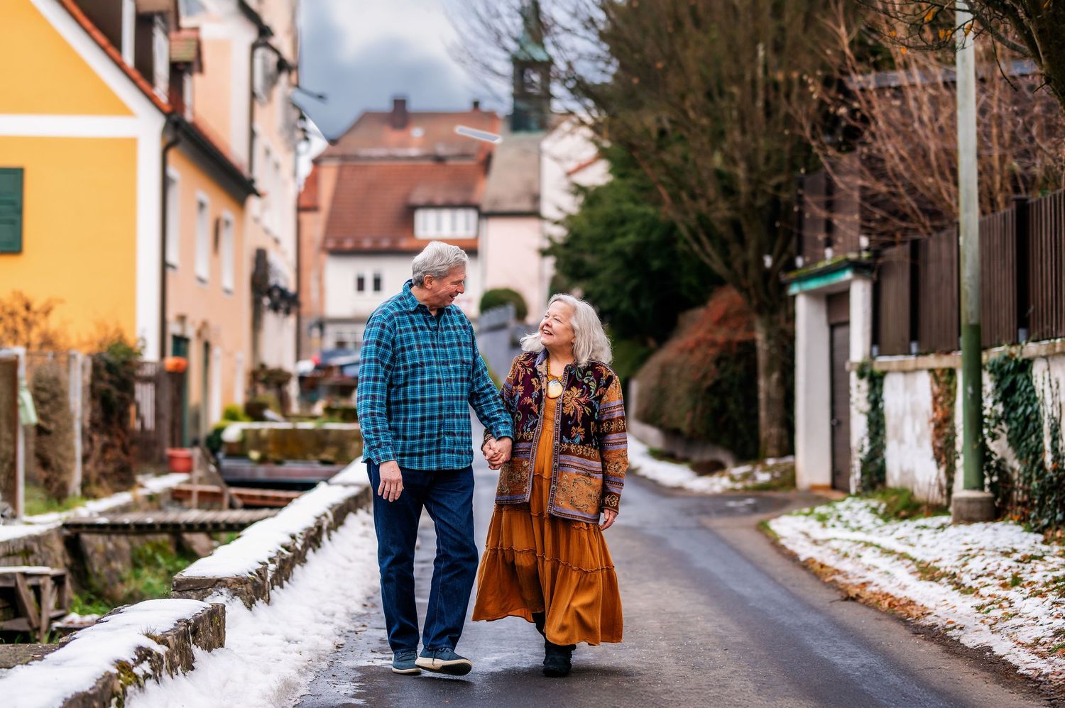 A couple walks together down a snowy village street lined with colorful houses in a quaint European town.