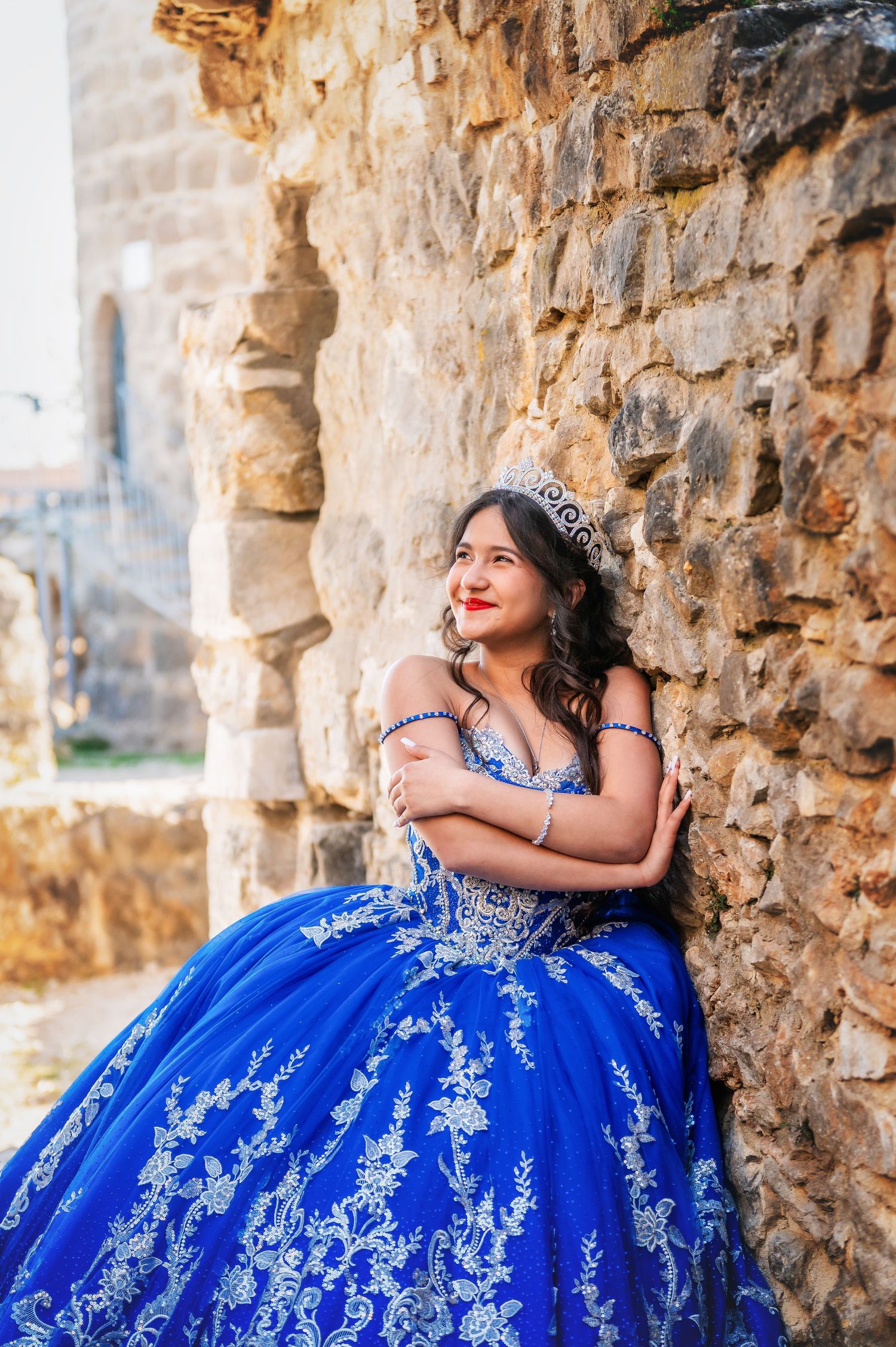 Young woman in a royal blue quinceañera ball gown with silver embroidery and tiara, leaning against a stone wall.