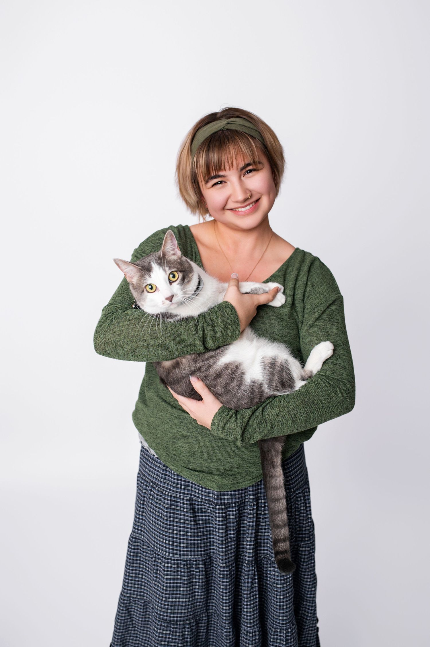 Smiling young woman in green sweater holding a gray and white tabby cat against white background.