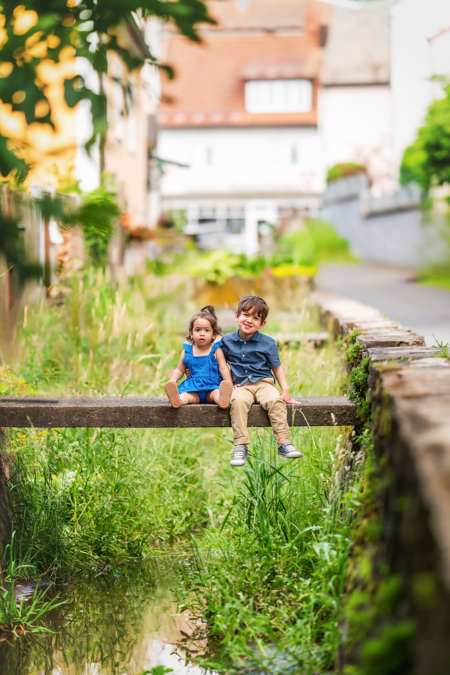 Two figures sitting together on a wooden bench surrounded by tall summer grass and greenery.