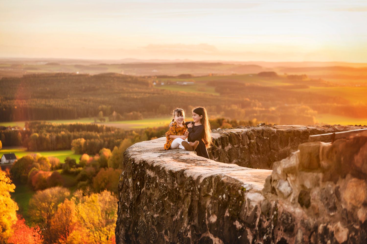 Two people sit on a rocky cliff edge enjoying a vibrant autumn sunset over rolling hills.