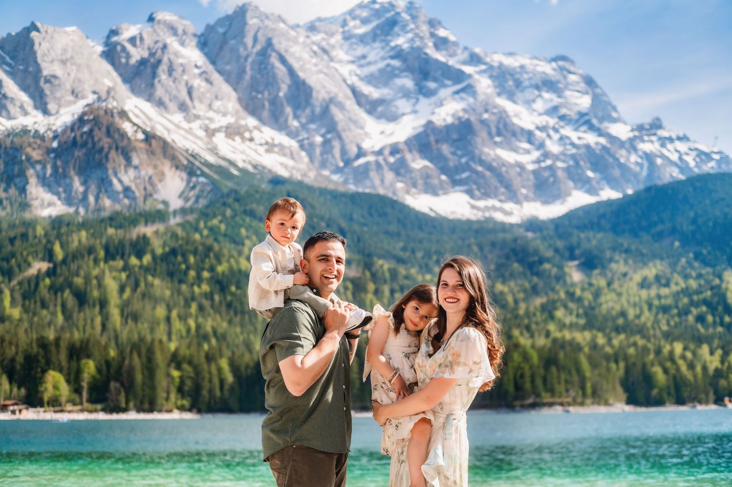Family of four posing by a turquoise alpine lake with snow-capped mountains and pine forests in the background.