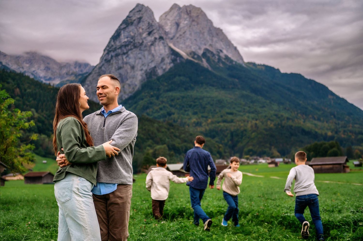 People enjoying a scenic moment in front of majestic mountain peaks in the Alps with green meadows and cloudy skies.