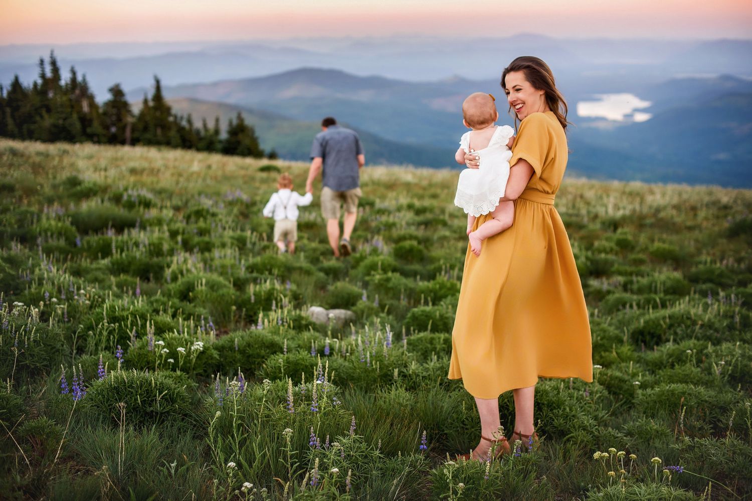 Family on mountain meadow at sunset, mother in yellow dress holding baby while father walks with dog.