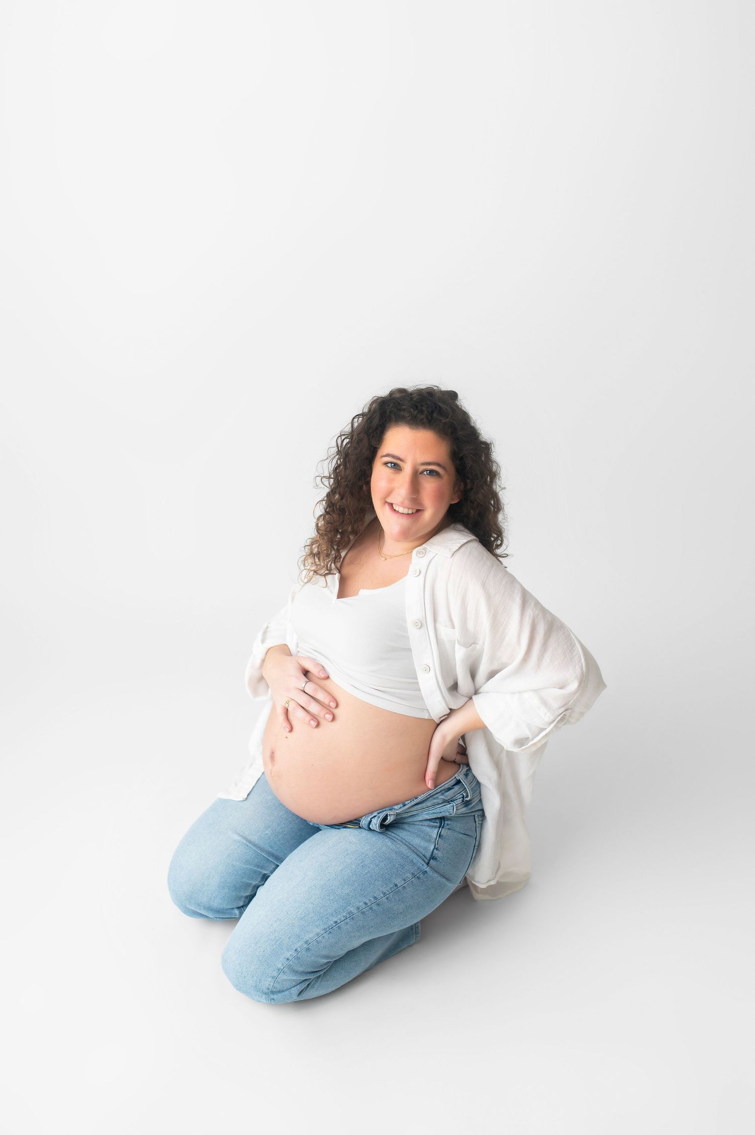 Smiling pregnant woman in white top and jeans sitting on floor cradling her baby bump against white background.