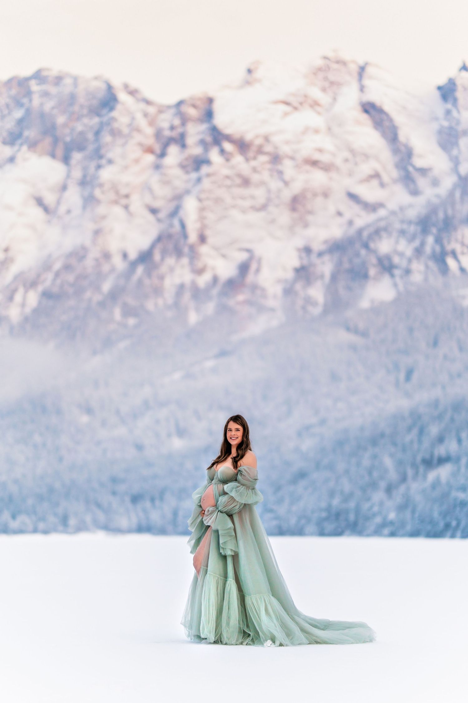 A flowing mint green gown creates an ethereal scene against snowy terrain and majestic mountain peaks at sunset.