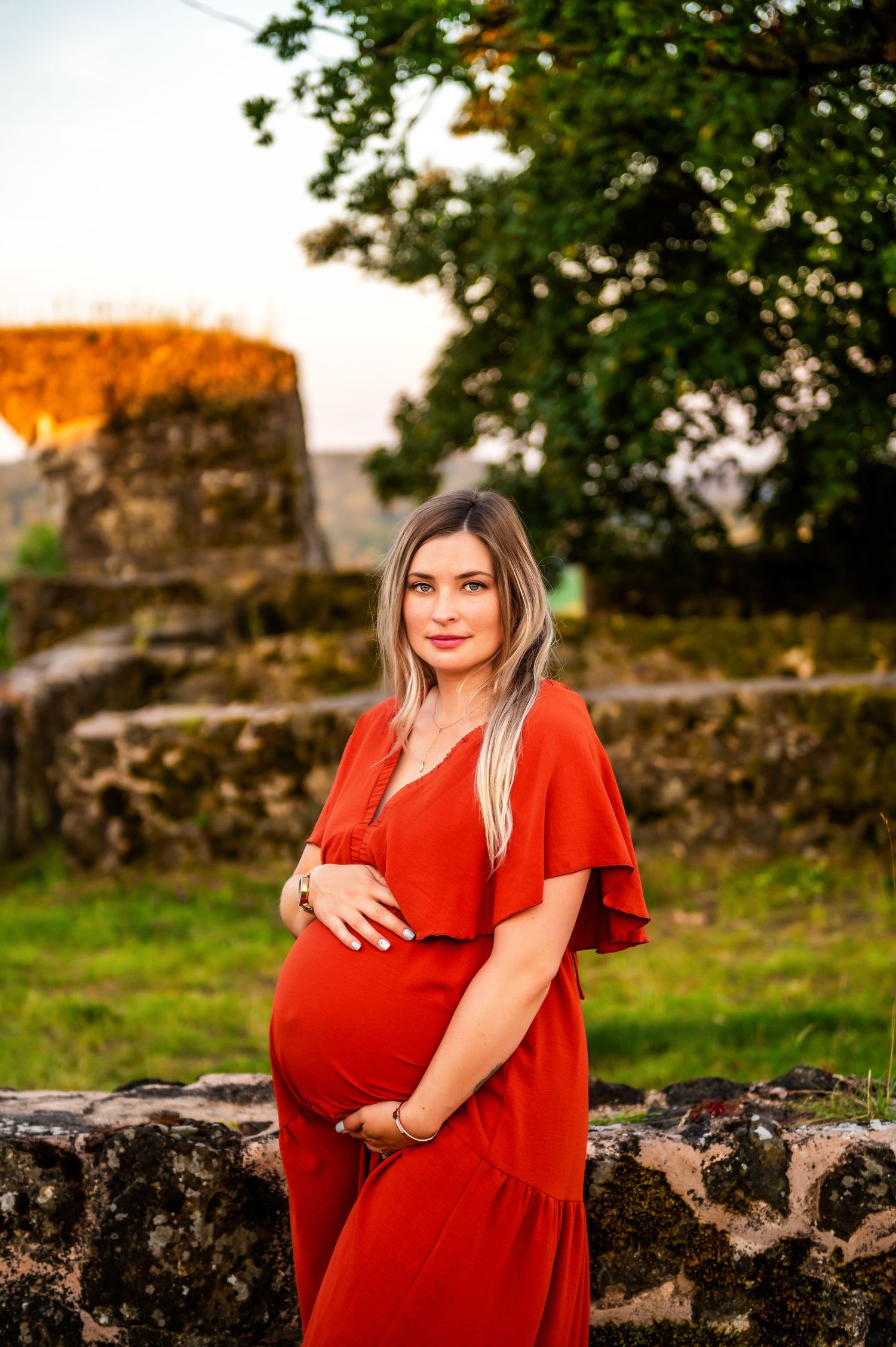 Expectant mother in flowing red dress poses against ancient stone ruins at sunset.