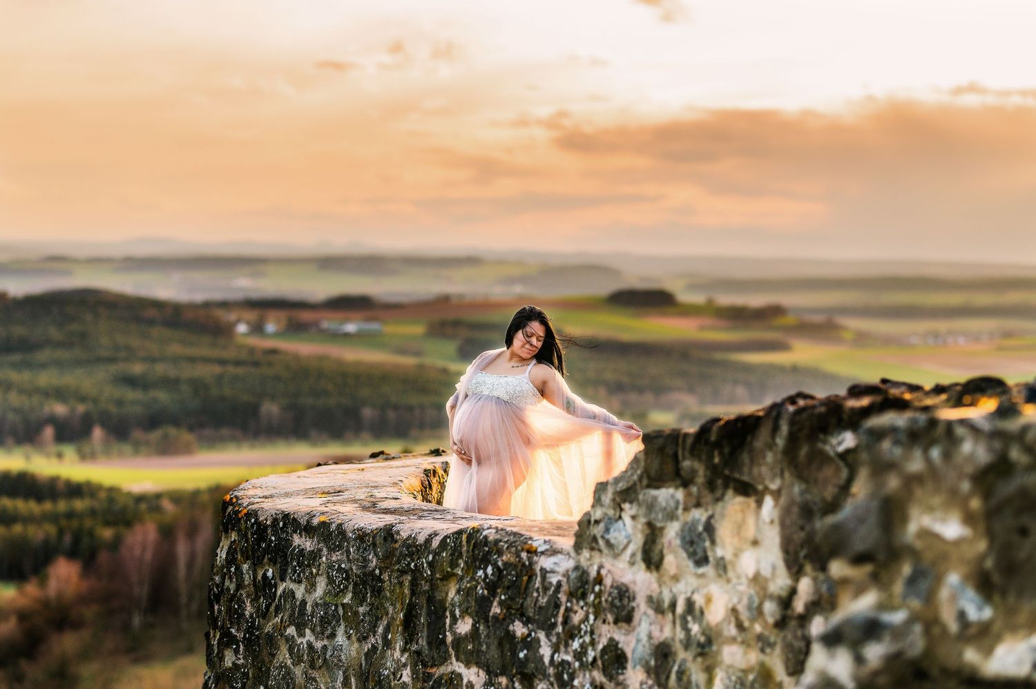 Maternity portrait on rocky cliff edge with dramatic sunset and rolling hills in background.