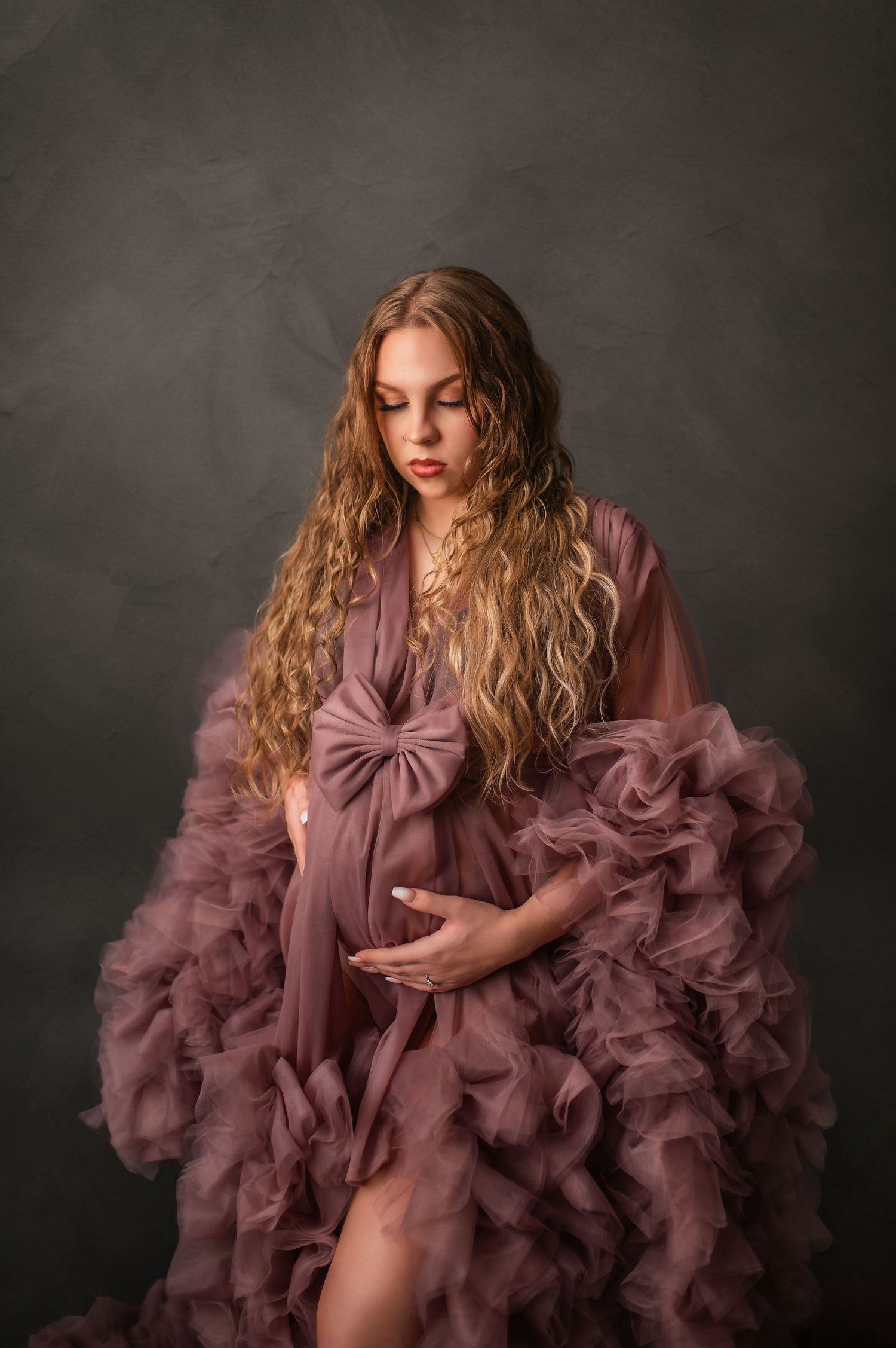 Dramatic maternity portrait featuring flowing mauve gown with ruffled details against dark backdrop.