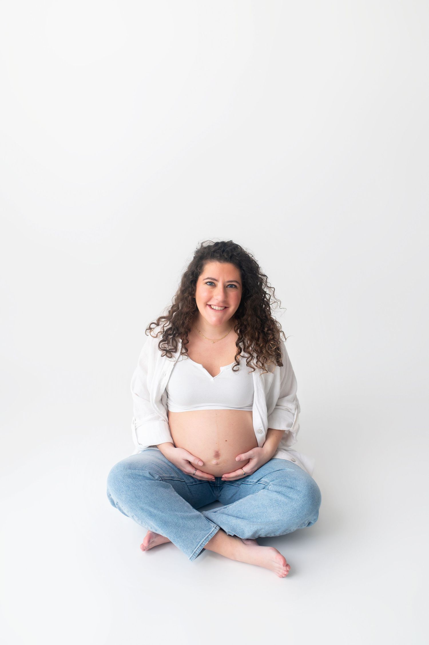 Expectant mother in white shirt and jeans poses cross-legged for maternity photos.