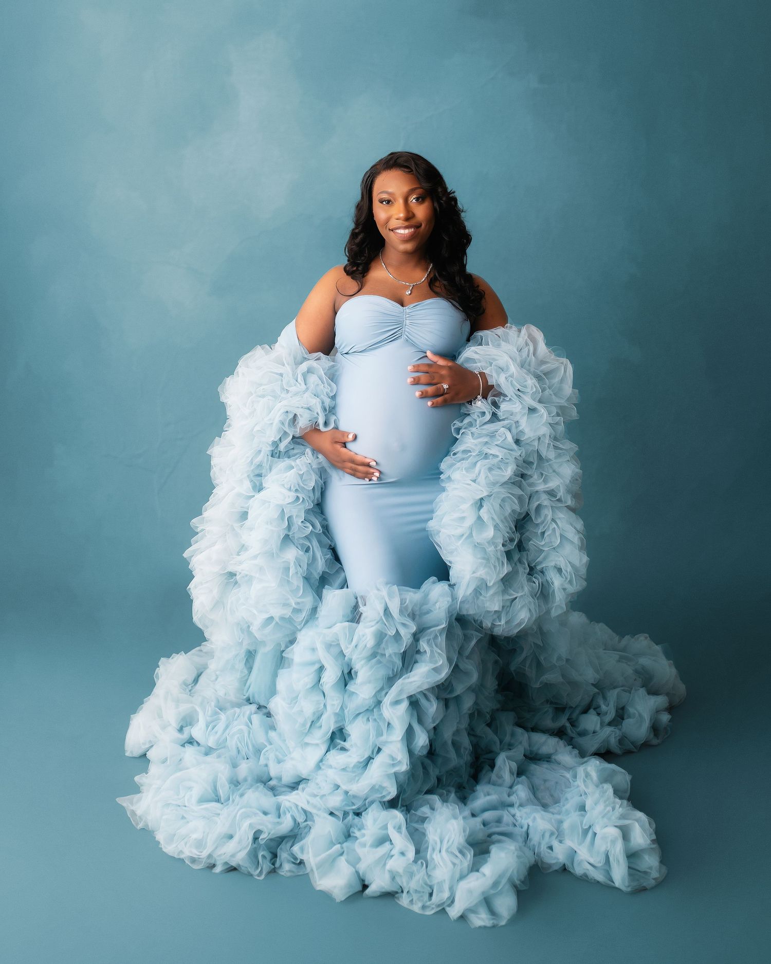 Maternity portrait featuring flowing blue ruffled dress against turquoise background.