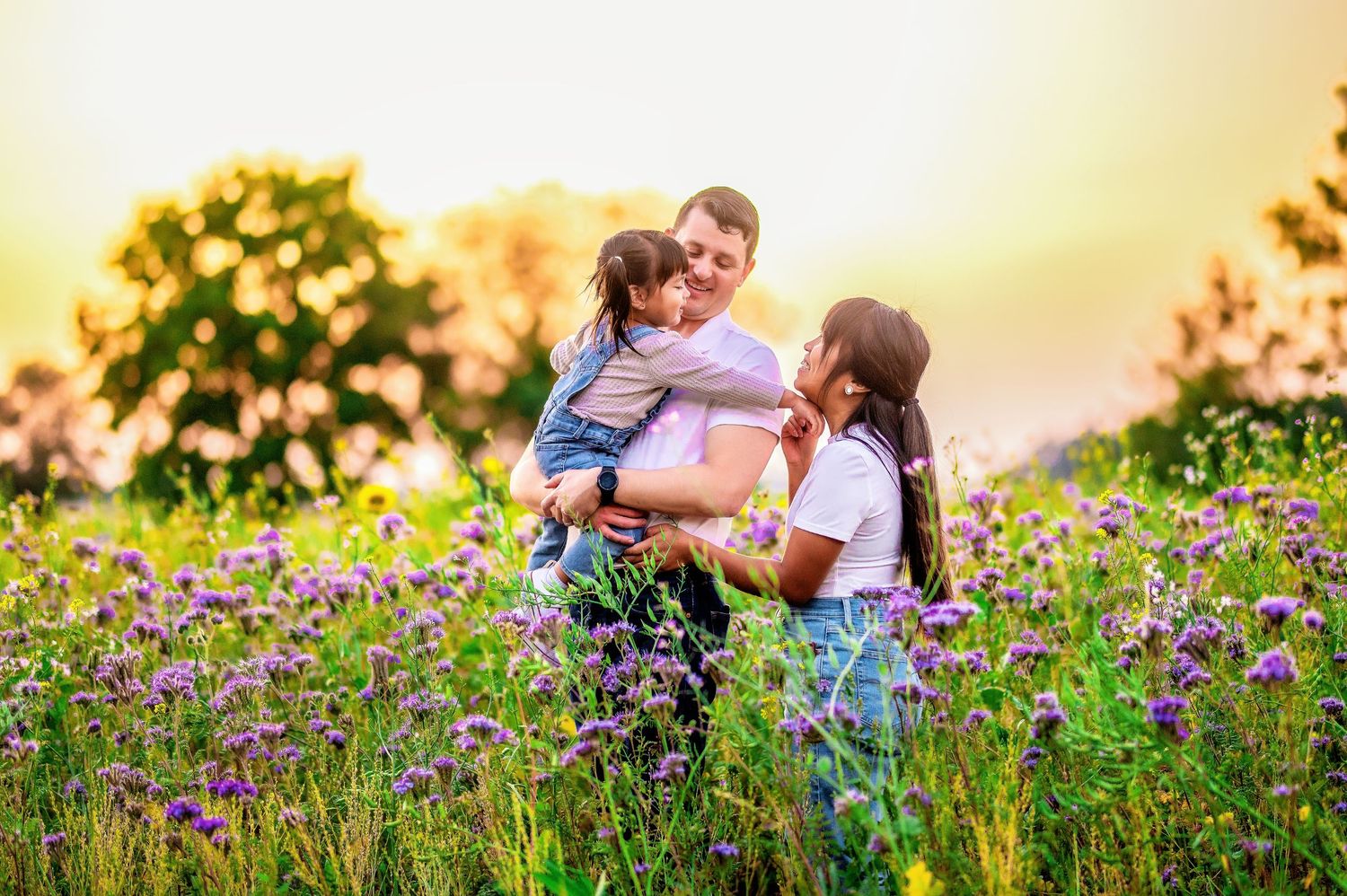 Parents embrace while playing with child in purple wildflower field during golden hour.