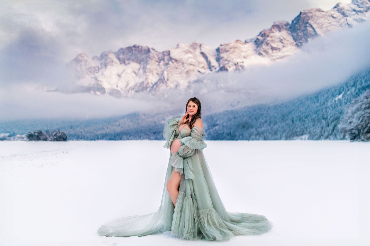 Woman in flowing sage green gown poses gracefully in snowy mountain landscape at sunset.