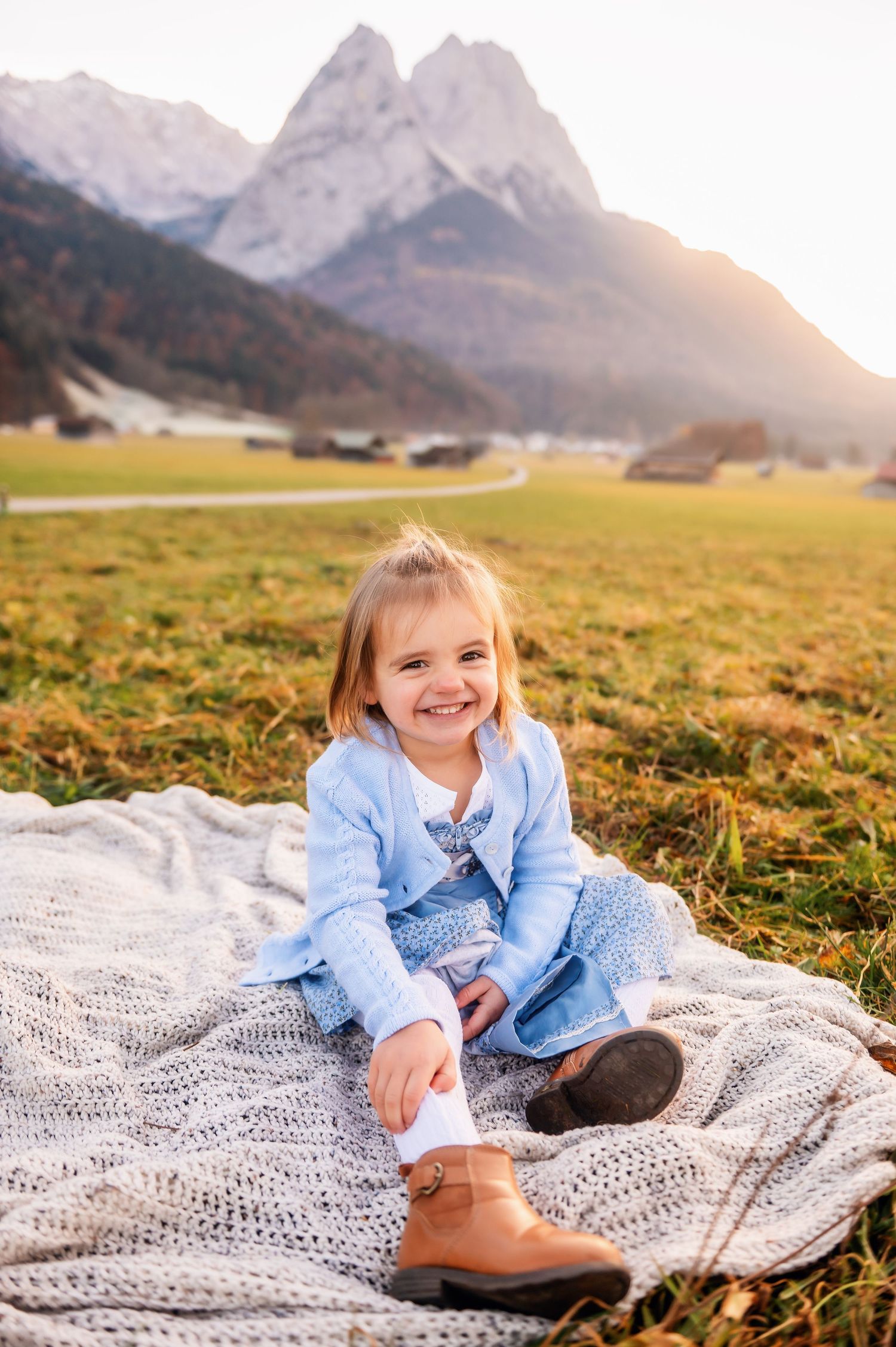 A child lies on a knitted blanket in a grassy meadow with majestic mountain peaks visible in the golden sunset.